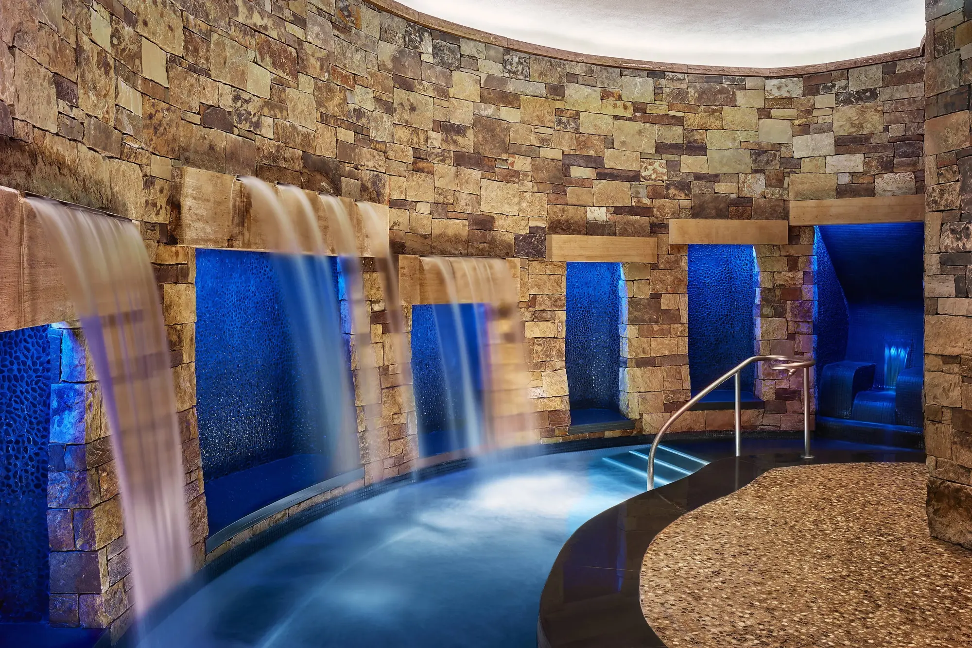 Cascading artificial waterfalls pour over a curved pool surrounded by stone walls and marble floors