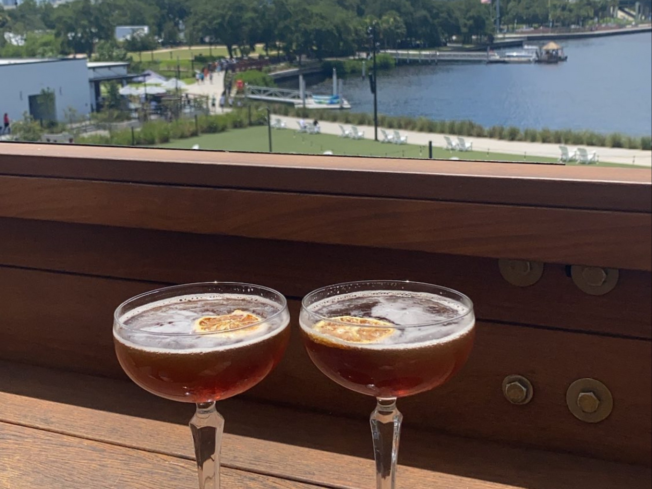 Two cocktails rest on a railing with a city skyline and water in the background under a blue sky.