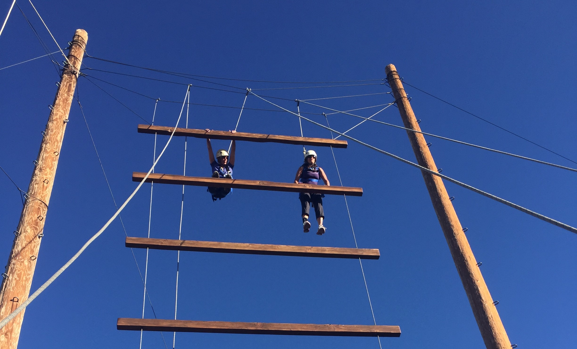 Giant hanging ladder on ropes with two people hanging, against a clear blue sky.