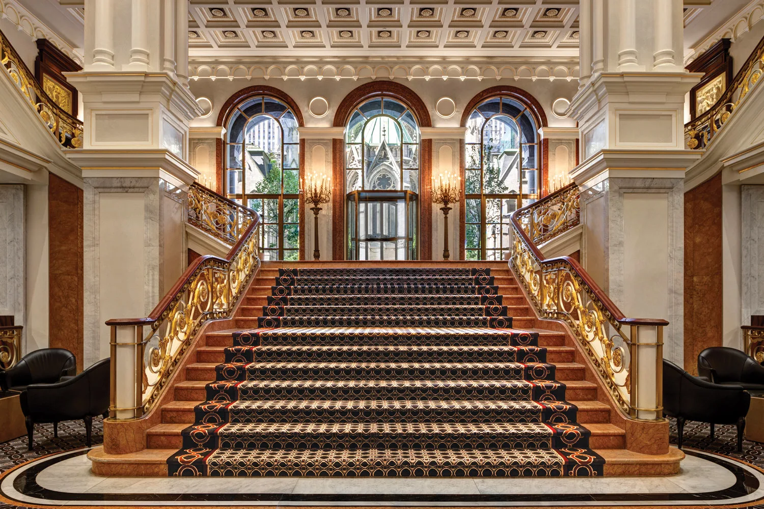 Elegant grand staircase with gilded railings, marble steps covered by a high-end rug leading up to glass doors and windows