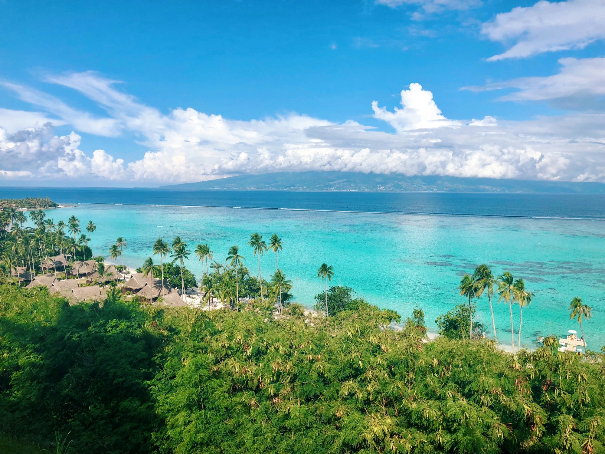 View of beach with palm trees, huts and crystal clear blue water under a partly cloudy sky.