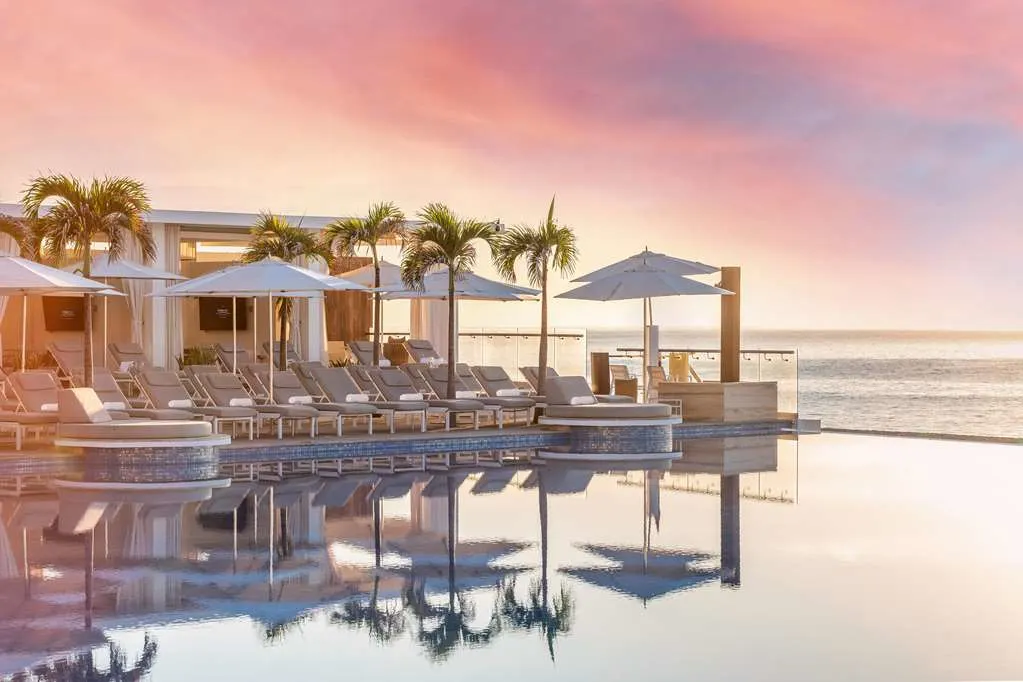 The edge of an infinity pool seems to blend into the ocean, while surrounded on one side by loungers and umbrellas underneath a pink sky