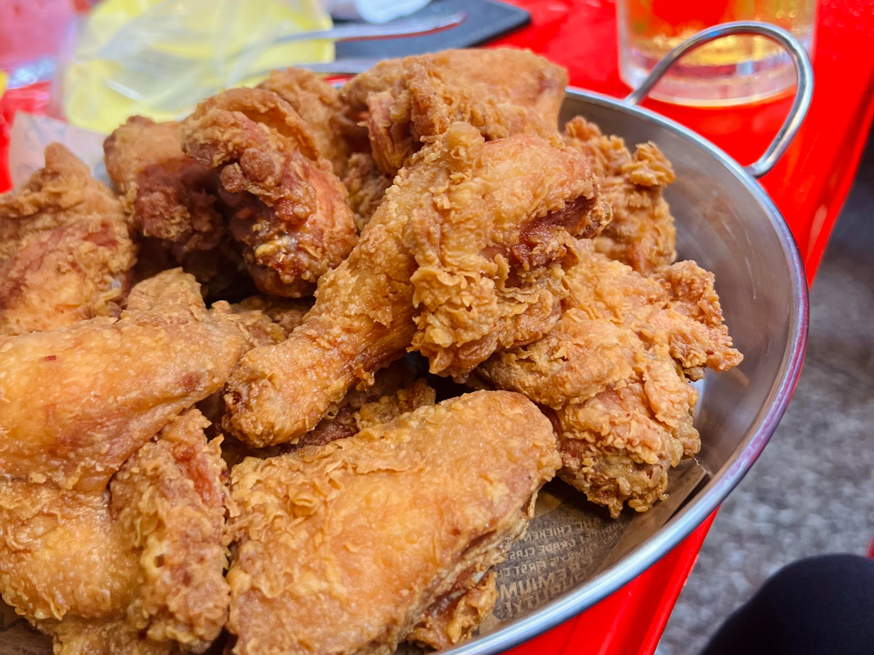 The image shows a bowl of fried chicken on a table with a glass of beer and plastic bags in the background