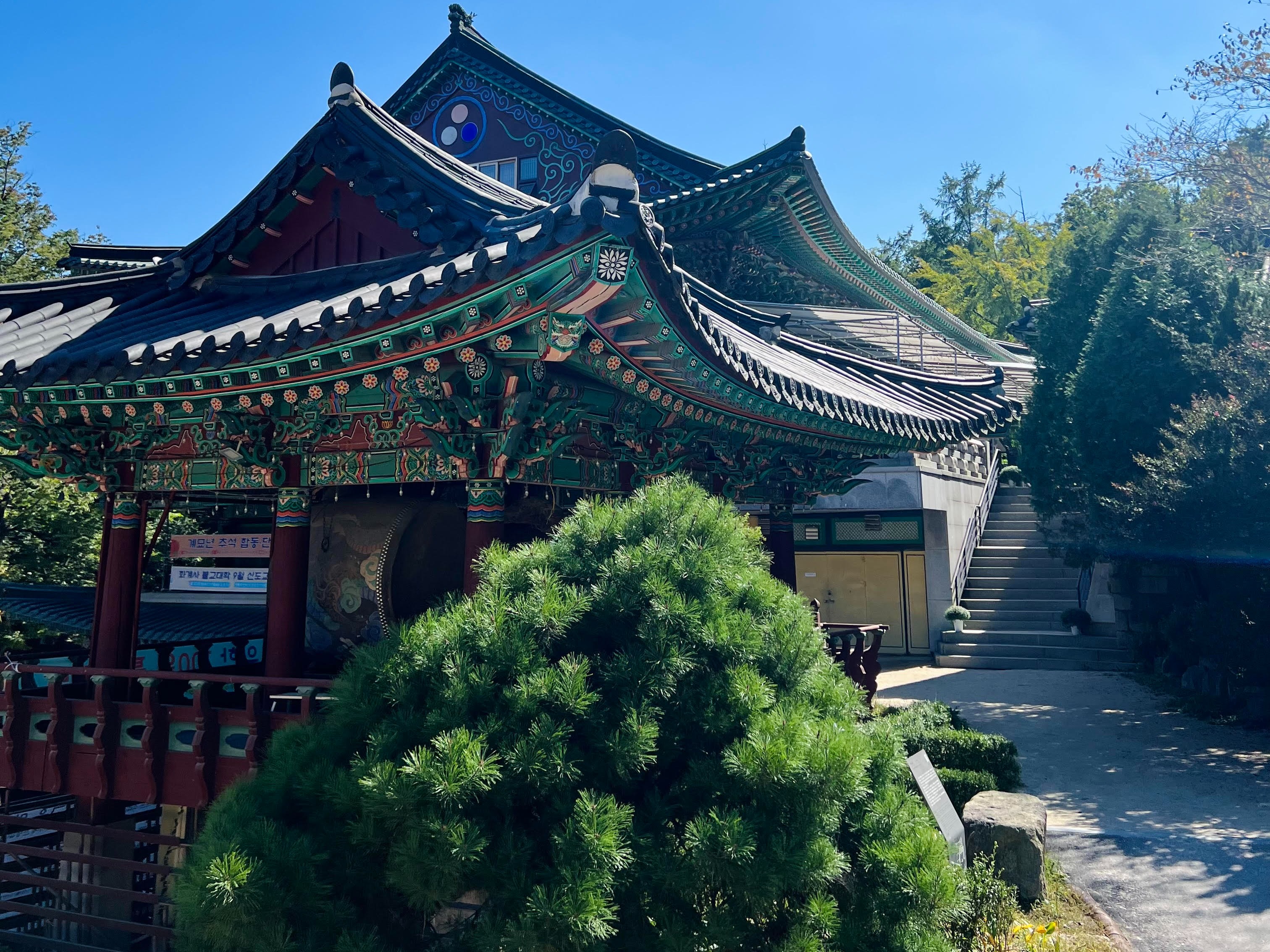 The image shows a traditional Korean-style building with intricate roof designs, set against a clear blue sky with sunlight streaming through.