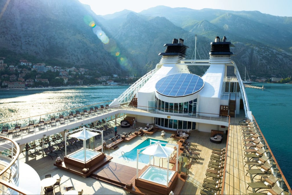 The top deck of a cruise ship with a chic pool area. In the distance, buildings of a town line the shores below mountains
