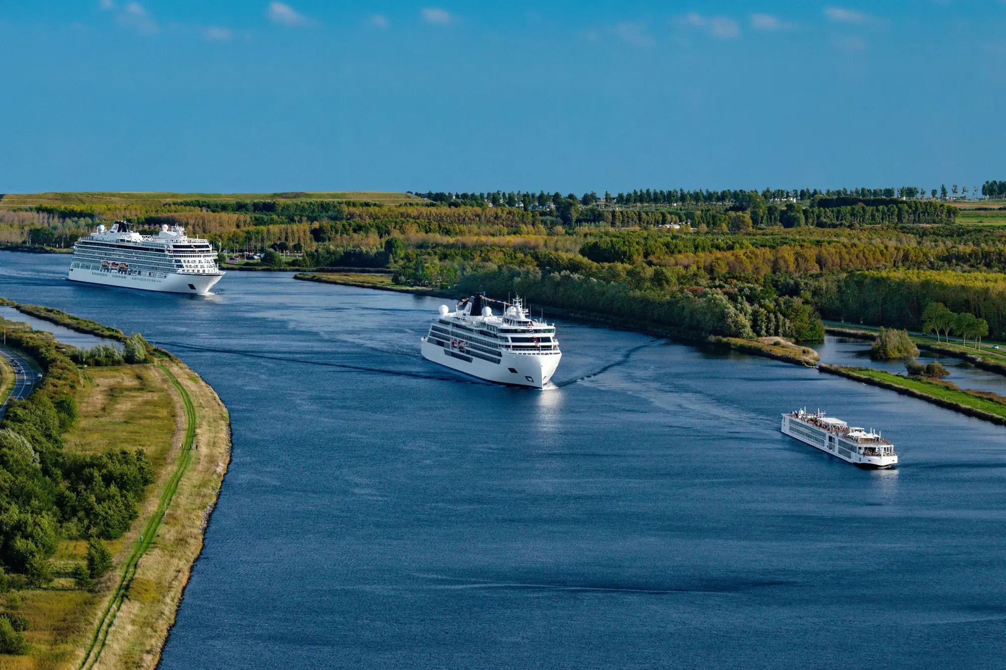 Three different Viking Cruise ships of varying sizes sail down a calm river surrounded by forests