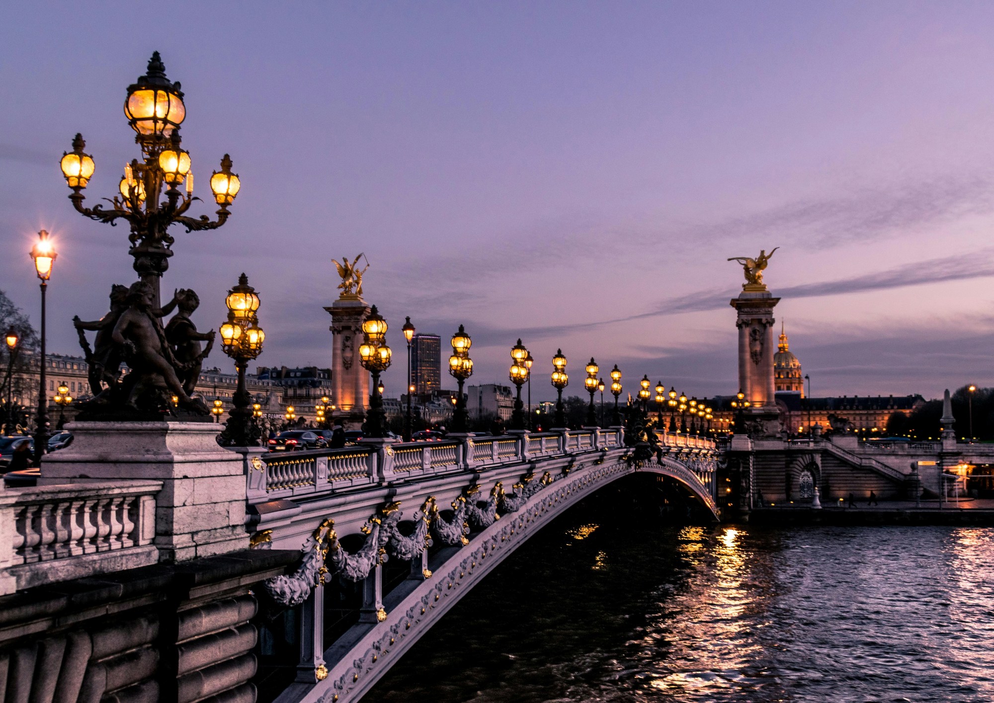 A bridge reaches across the Seine at dusk dotted with lit street lamps as water gently laps below.