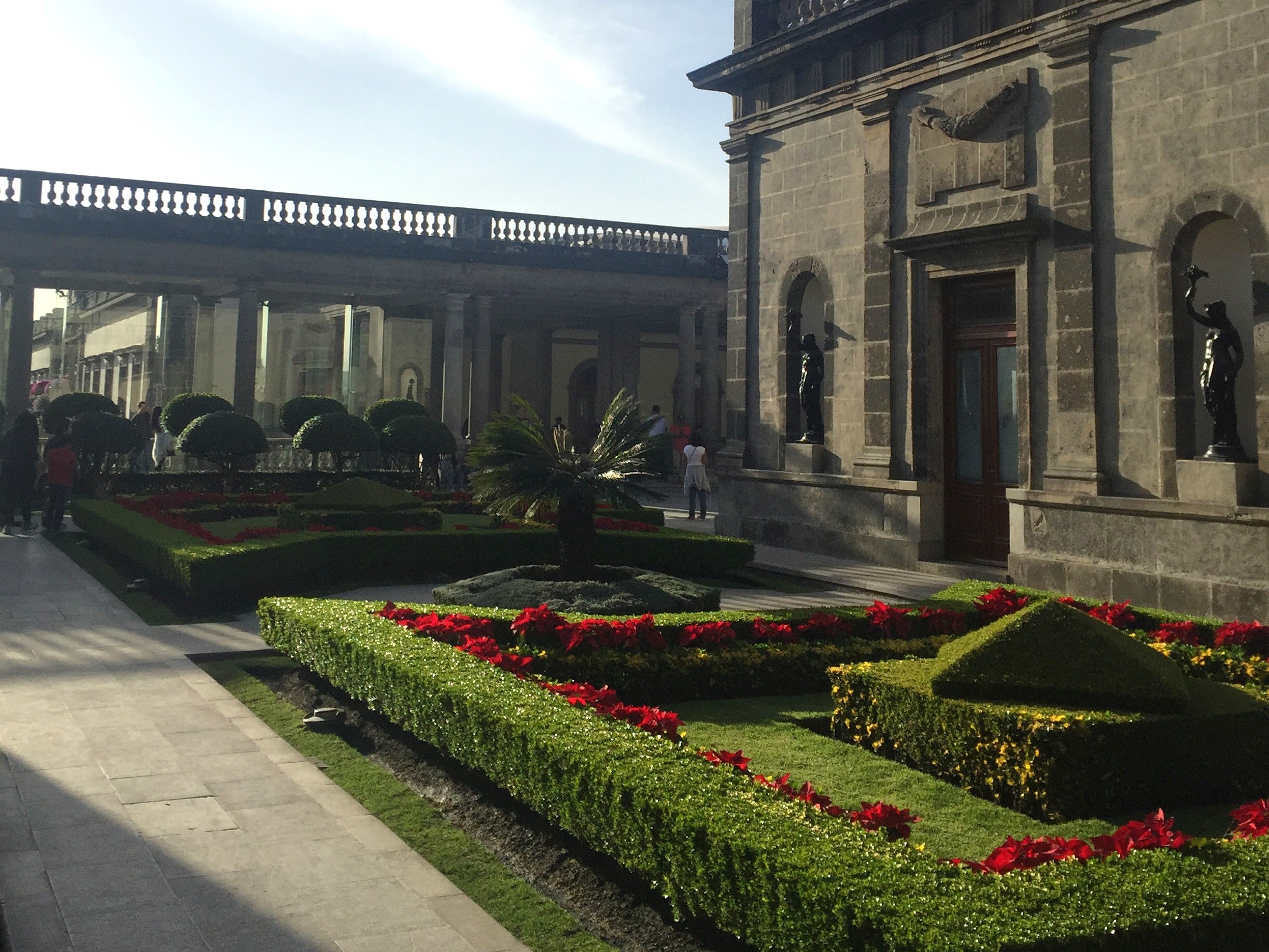 A serene garden with geometric hedges and vibrant flowers in front of an ornate building with a tower.