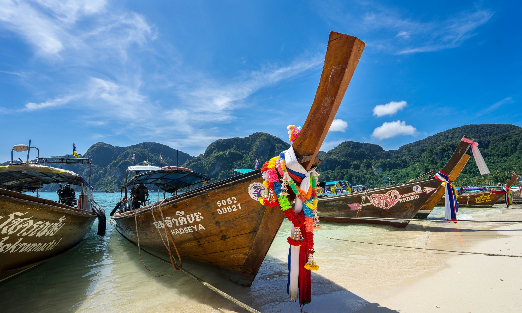 A line of island long tail boats dock against the sand as waves gently lap the shore on a clear day.
