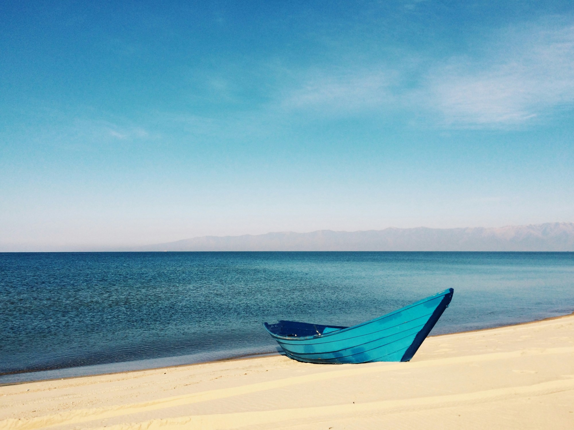 A boat is docked on the sand of a deserted beach as waves gently lap the shore on a sunny day.
