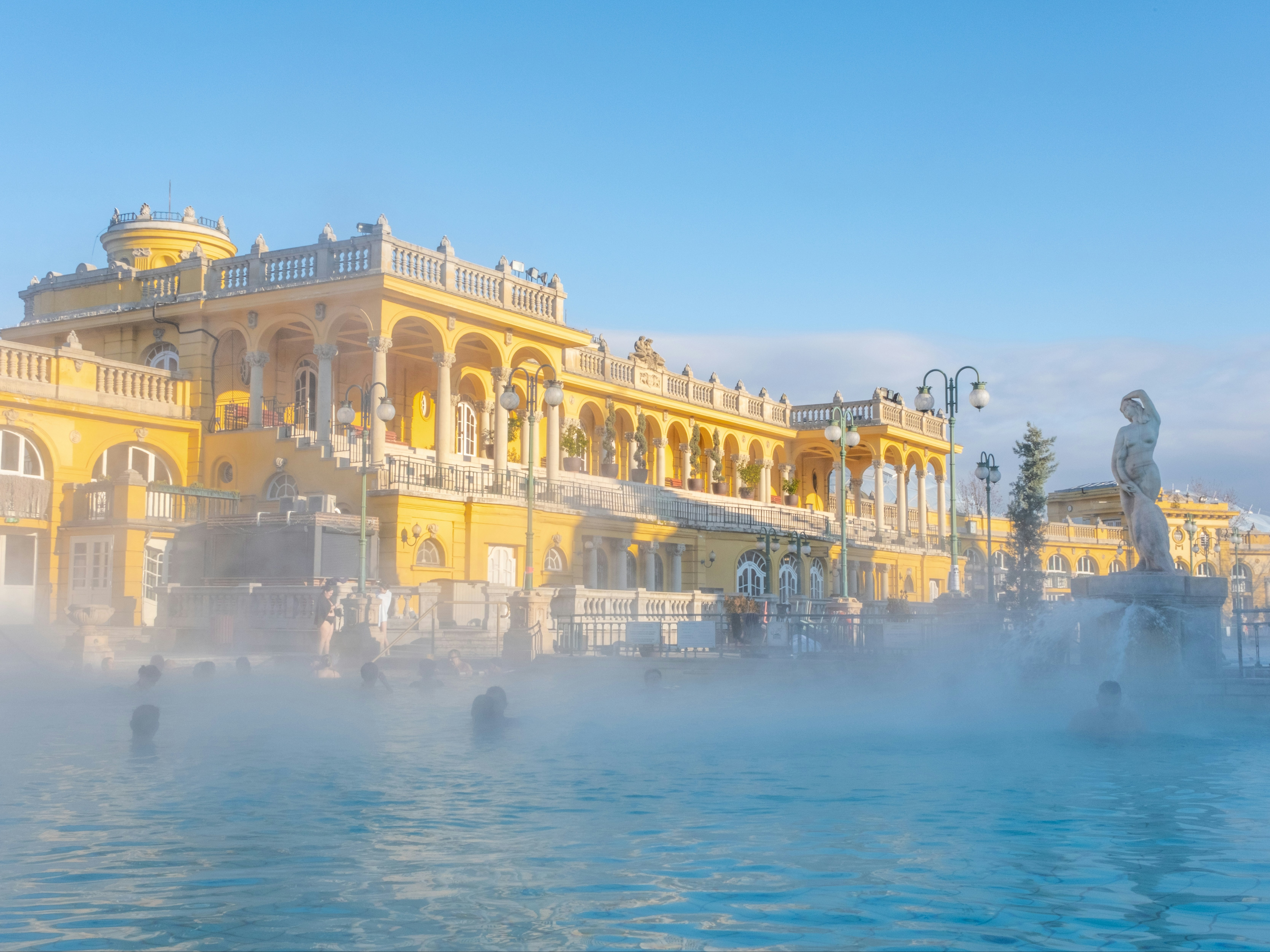 This image shows an outdoor thermal bath with people enjoying the water, set against an ornate, yellow building with domes.