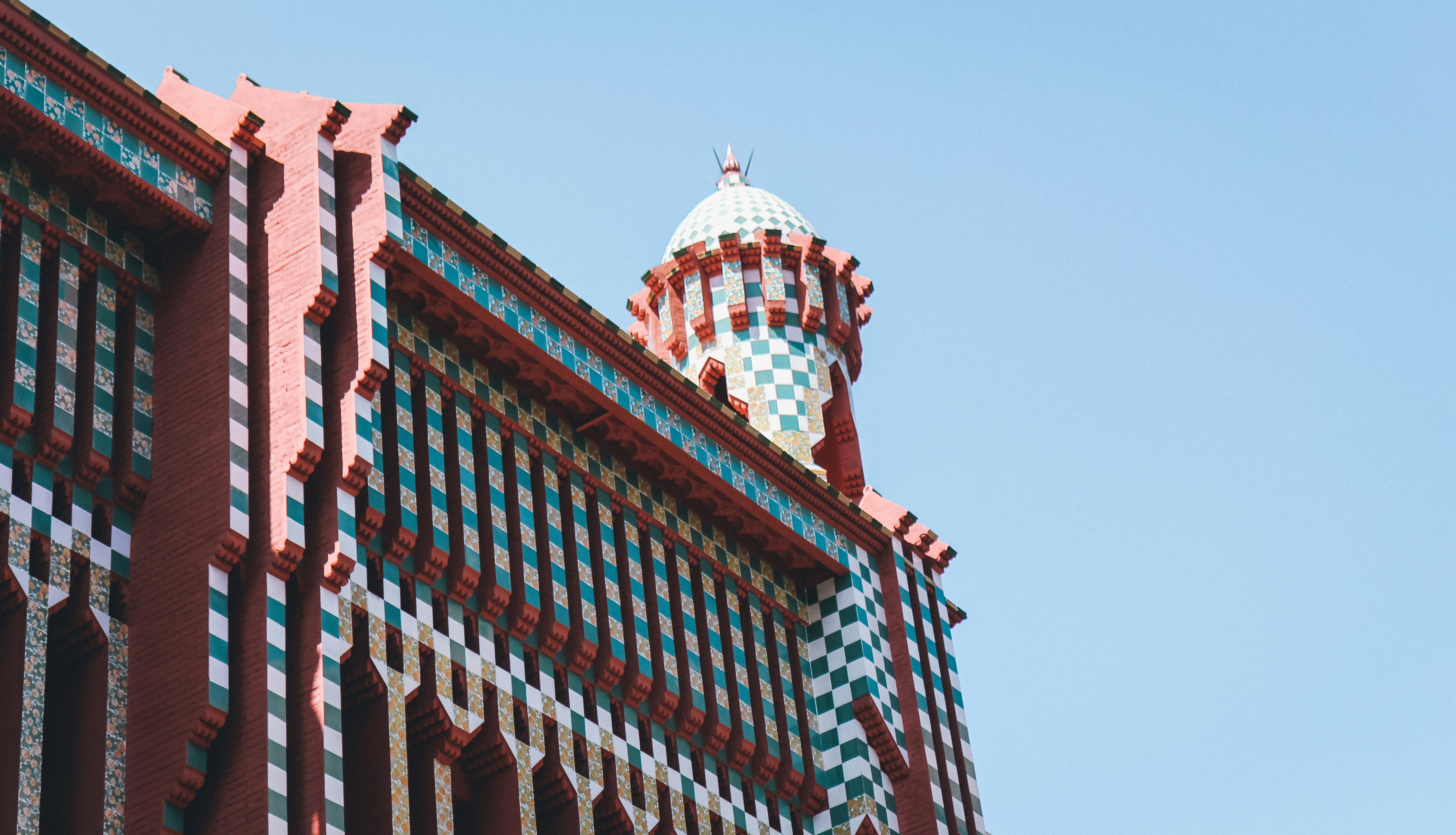 A view of the top of Casa Vicens in Barcelona with red, blue and white mosaic tiles and a turret, against blue sky.