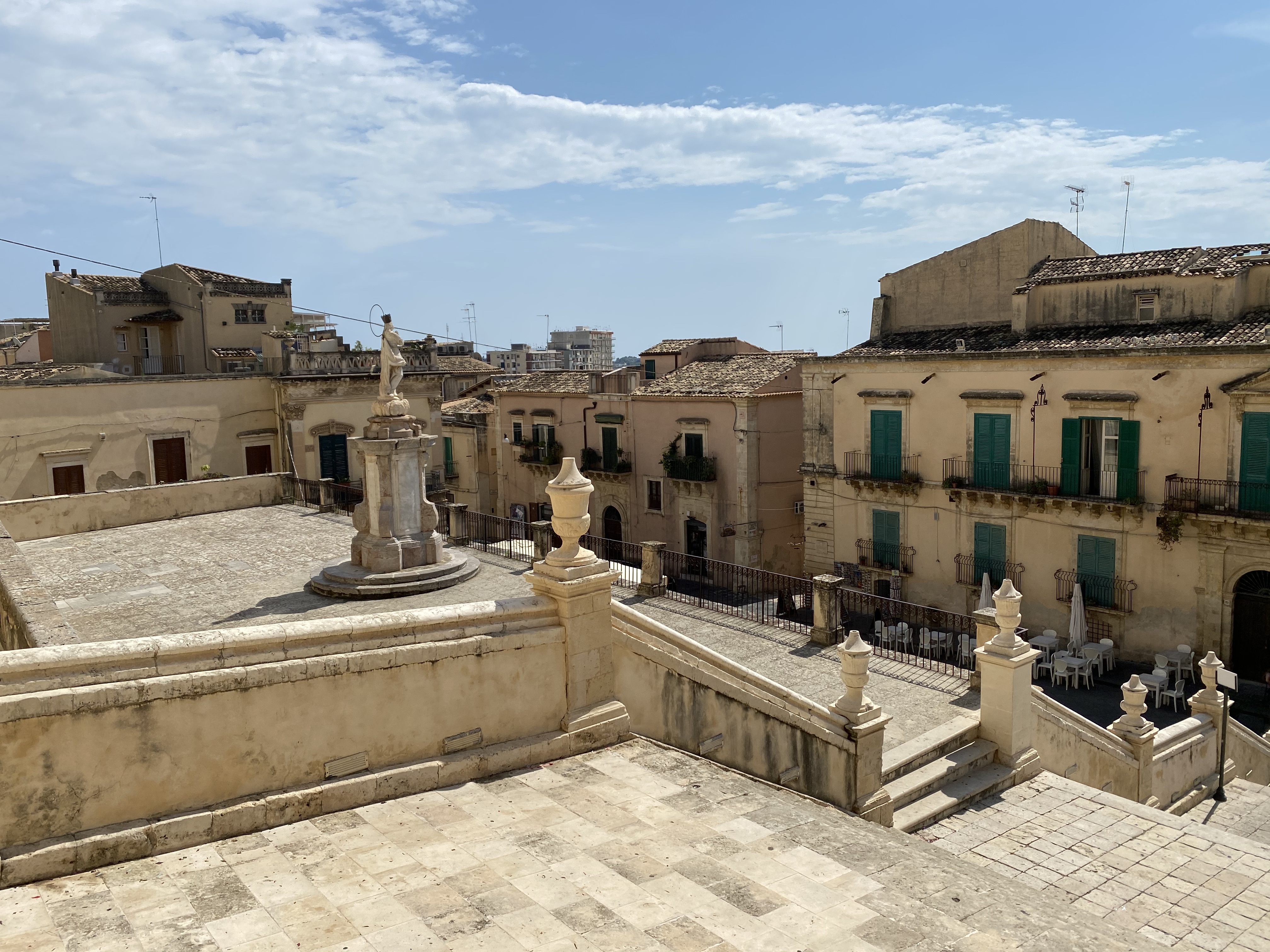 A view of Noto, Sicily showing a square with steps and a statue in the middle with surrounding old budilings.