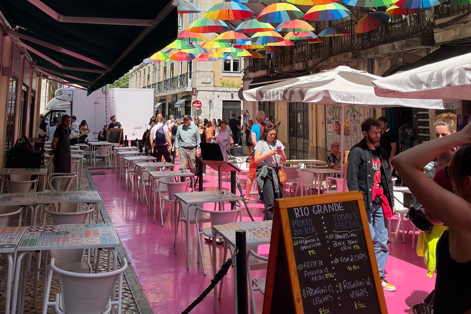 A pink-painted street with an outside dining area and rainbow-colored umbrellas above