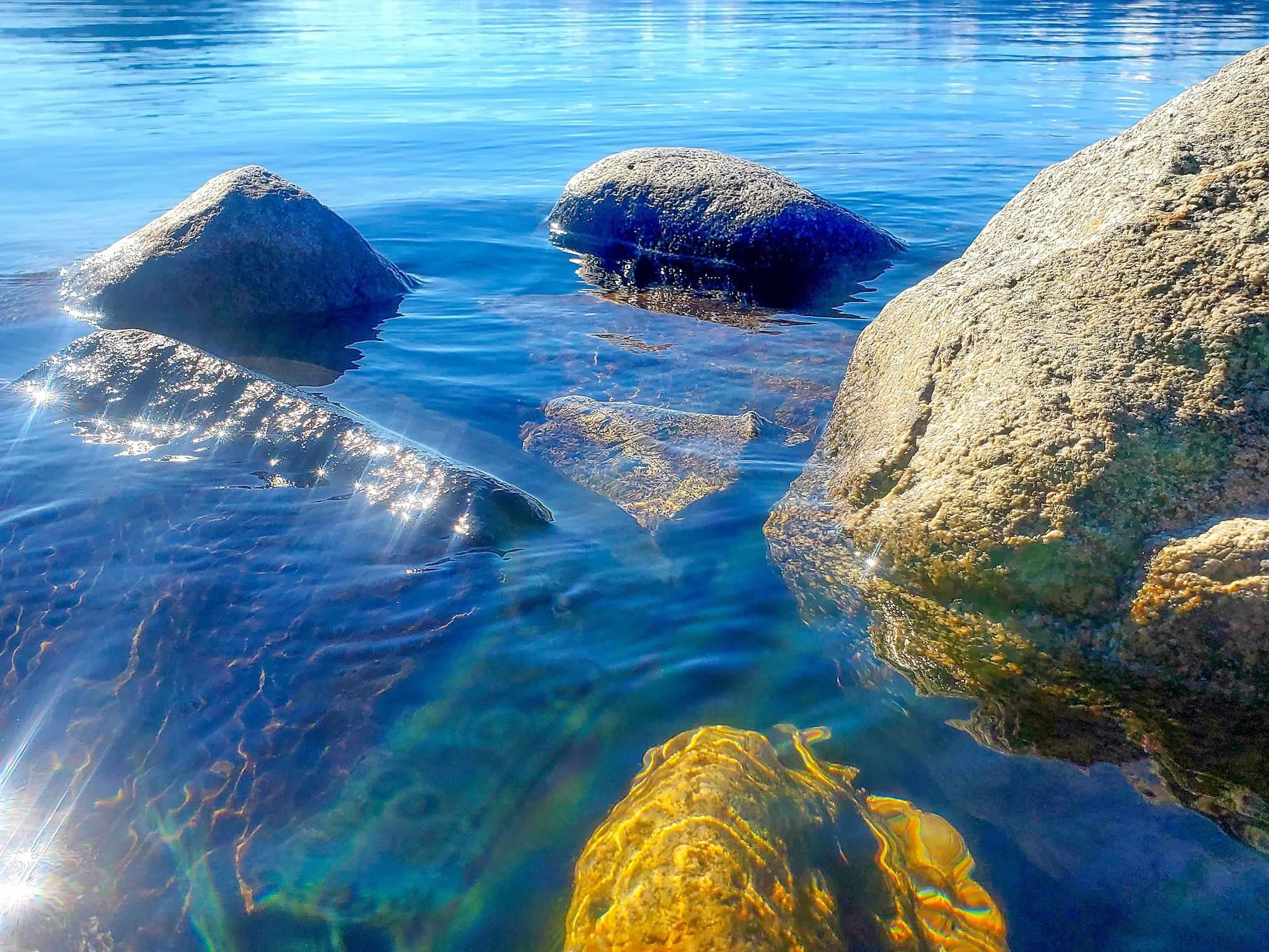 A clear lake with submerged rocks and mountains in the background under a blue sky.
