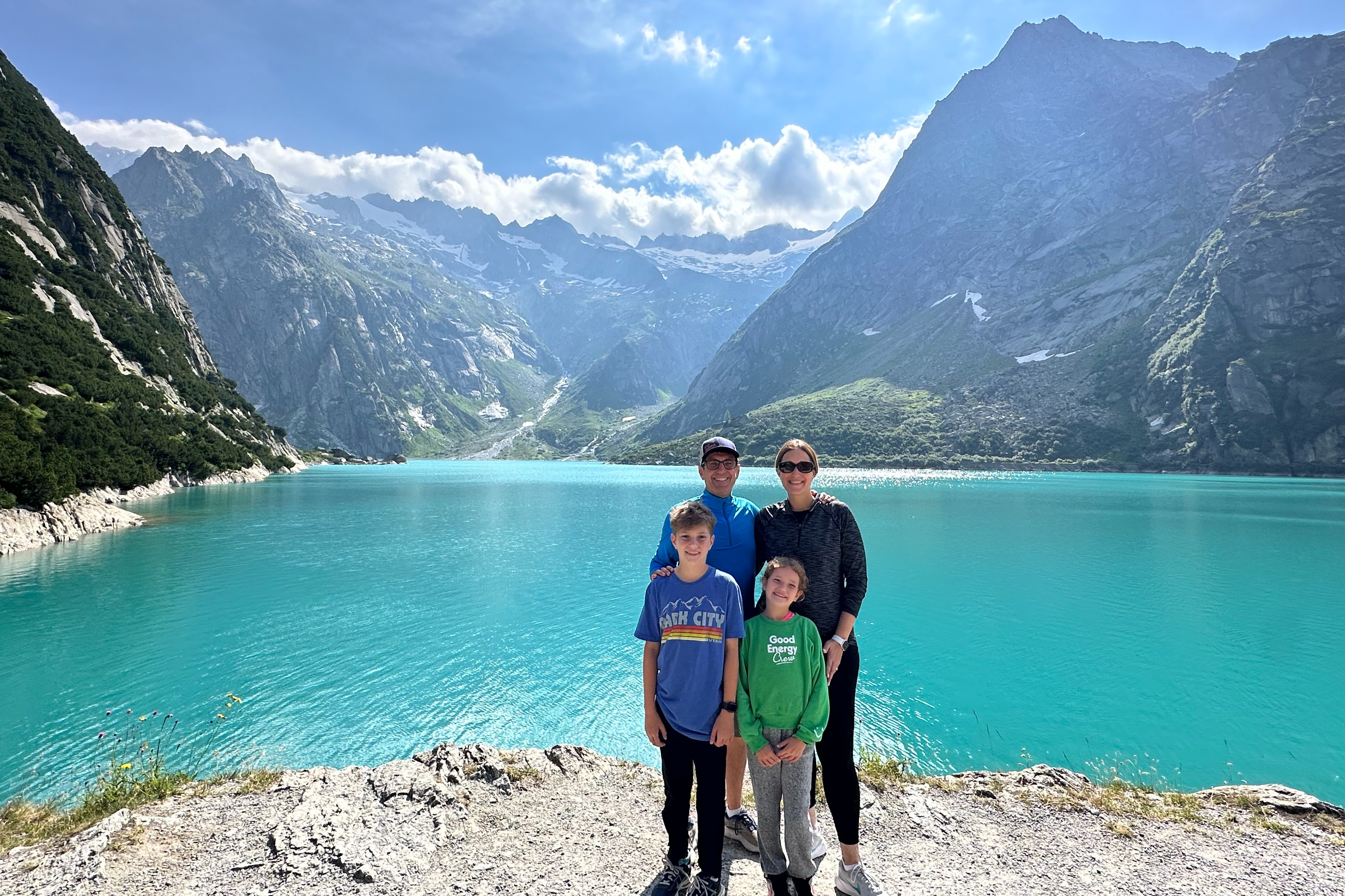 Family of four posed in front of a blue alpine lake with mountains in the background.