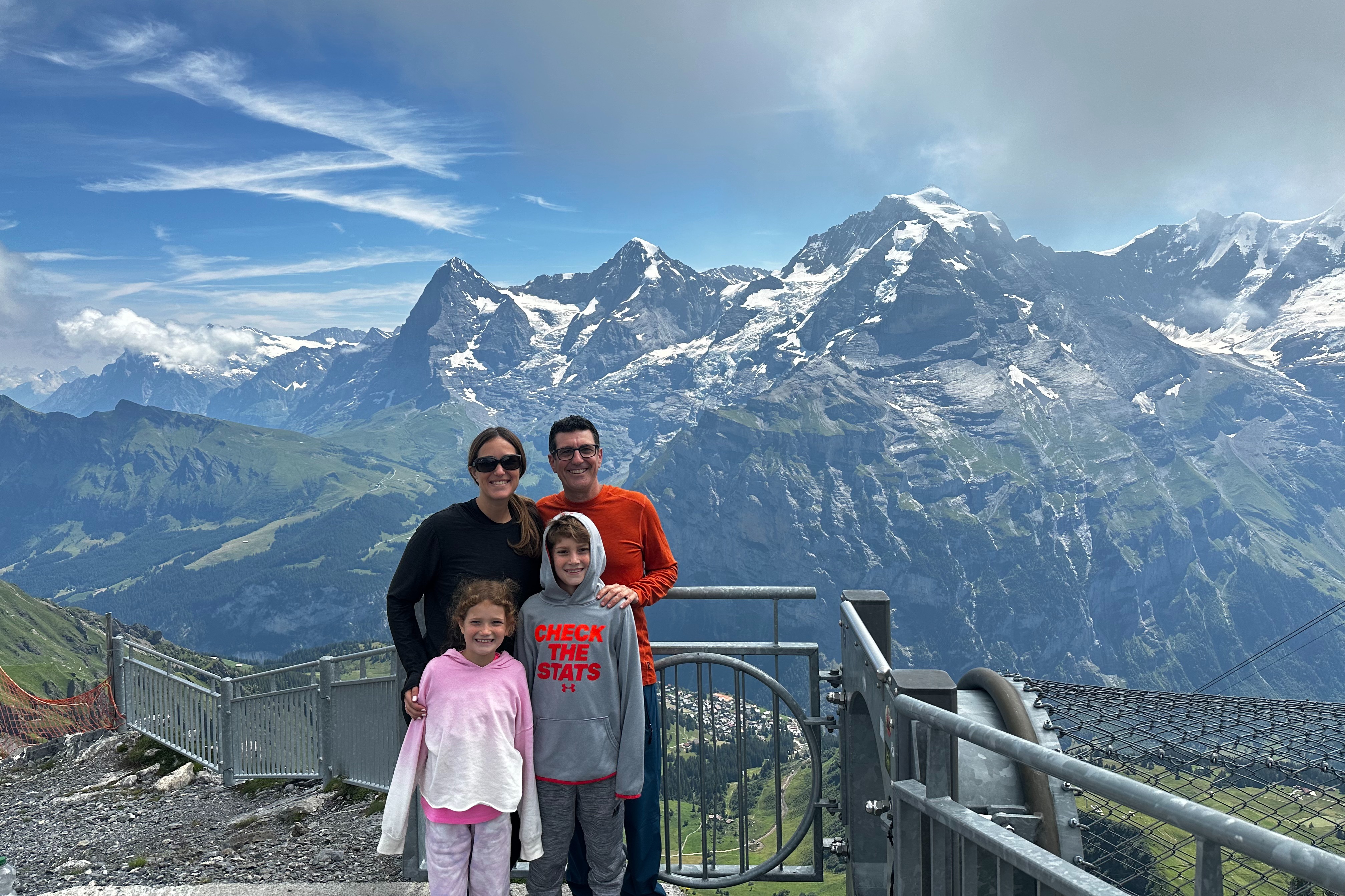 A family of four poses on a mountain top with snowy peaks behind.
