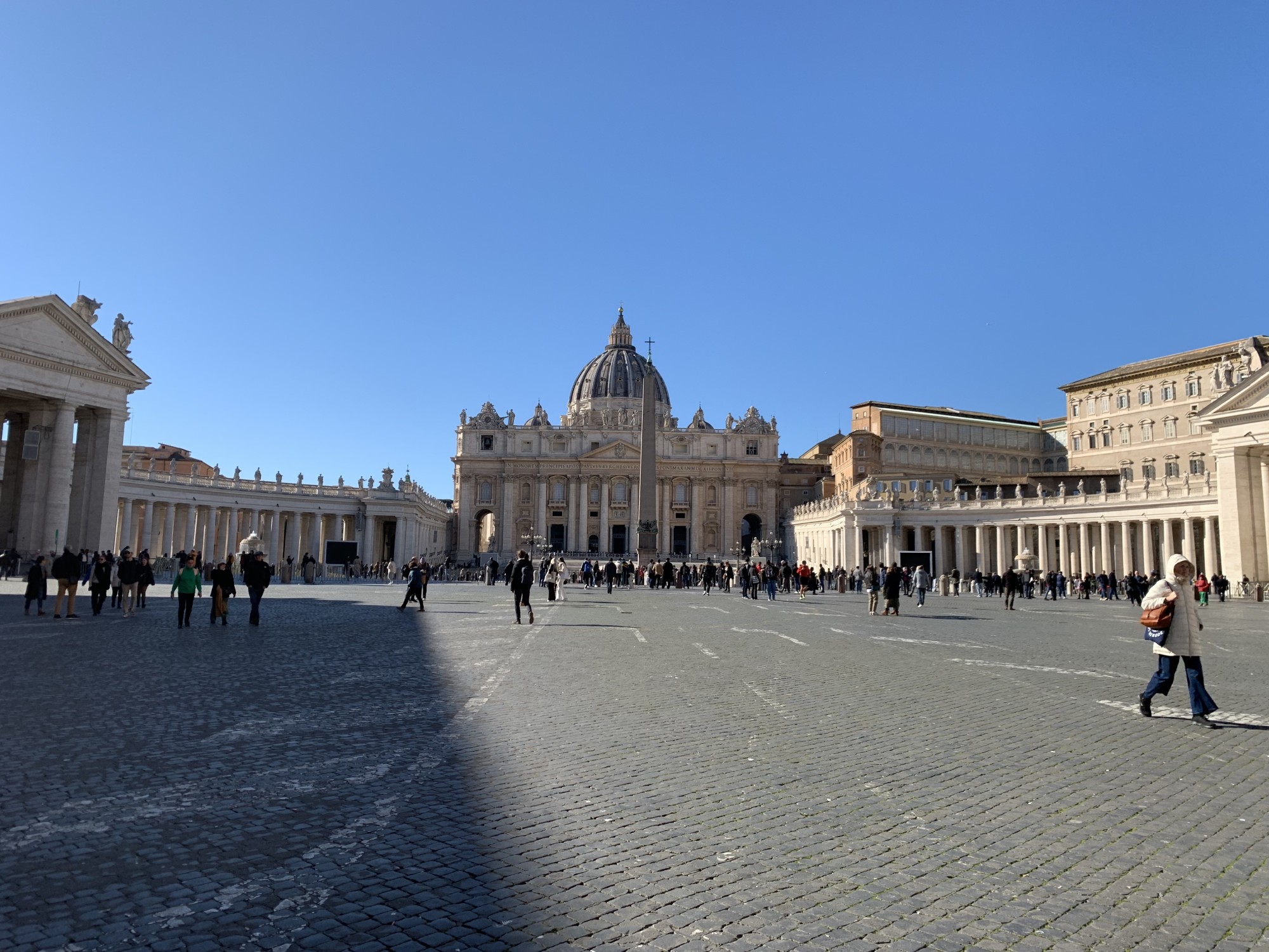 People walking in a square surrounded by buildings in the morning light on a clear day.