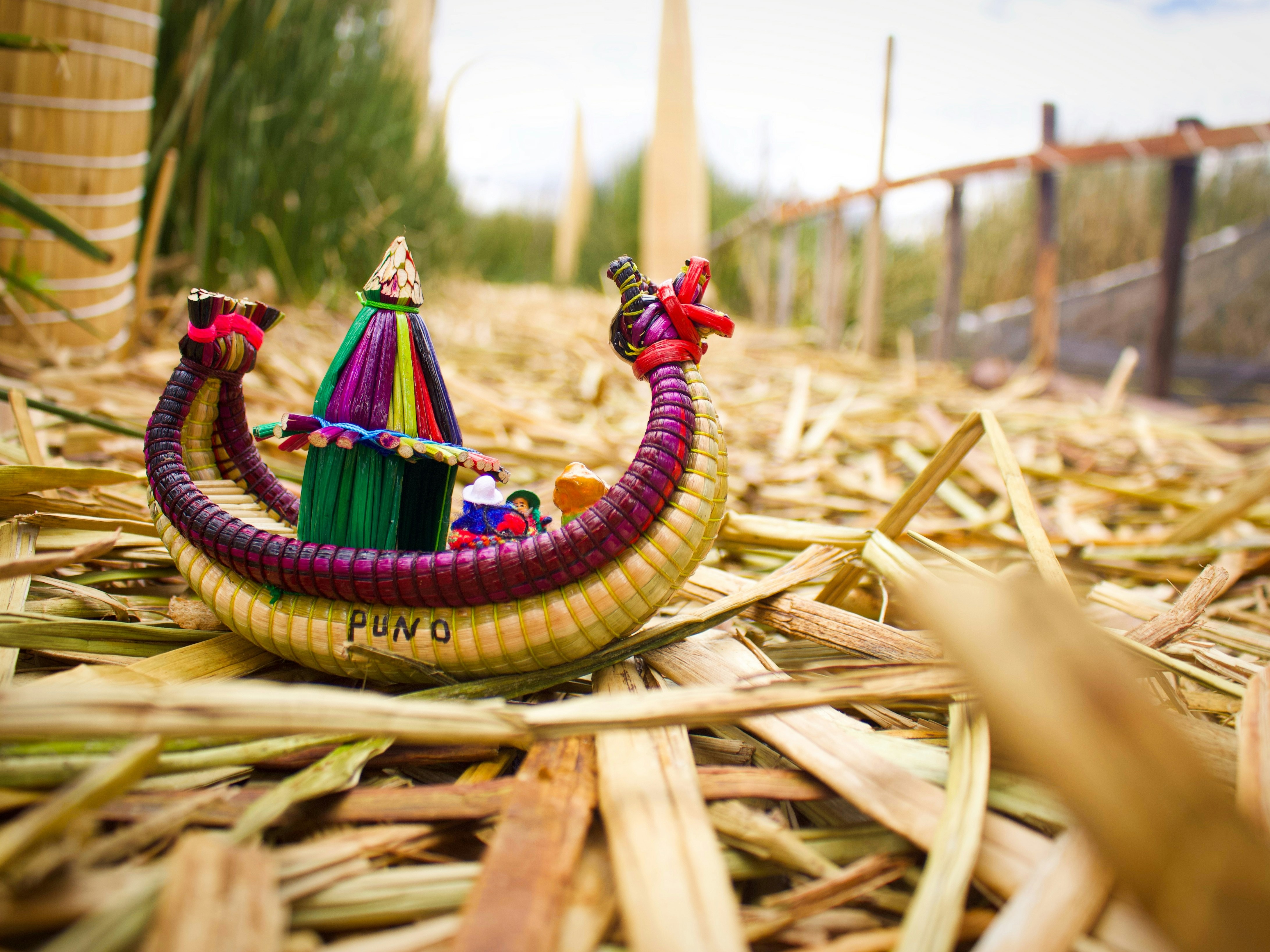 A colorful miniature reed boat rests on a bed of dried reeds with “PUNO” written on the side, showcasing traditional craftwork.