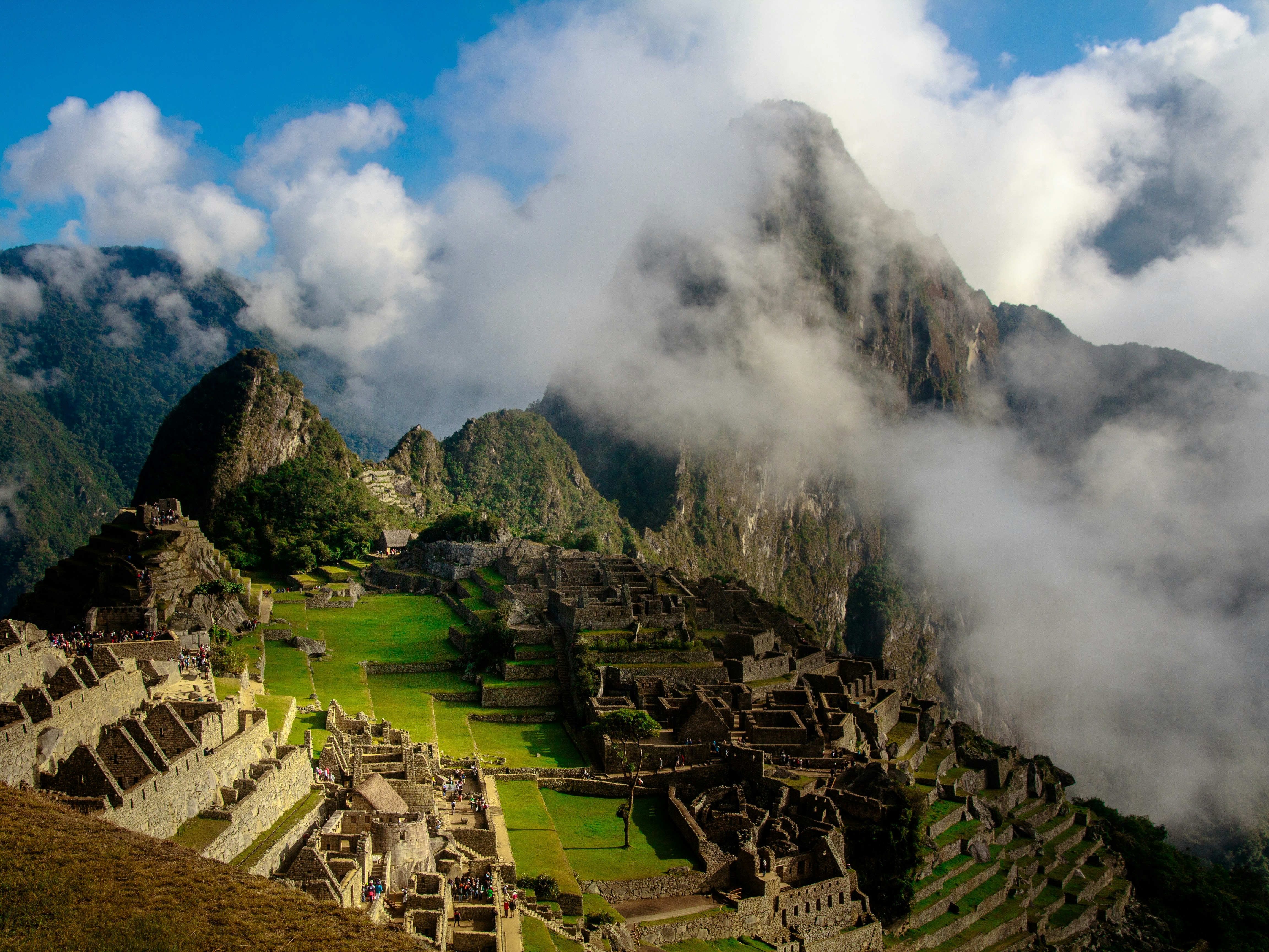 The ancient Incan city of Machu Picchu with its iconic ruins and terraces is nestled between mountain peaks under a partly cloudy sky.