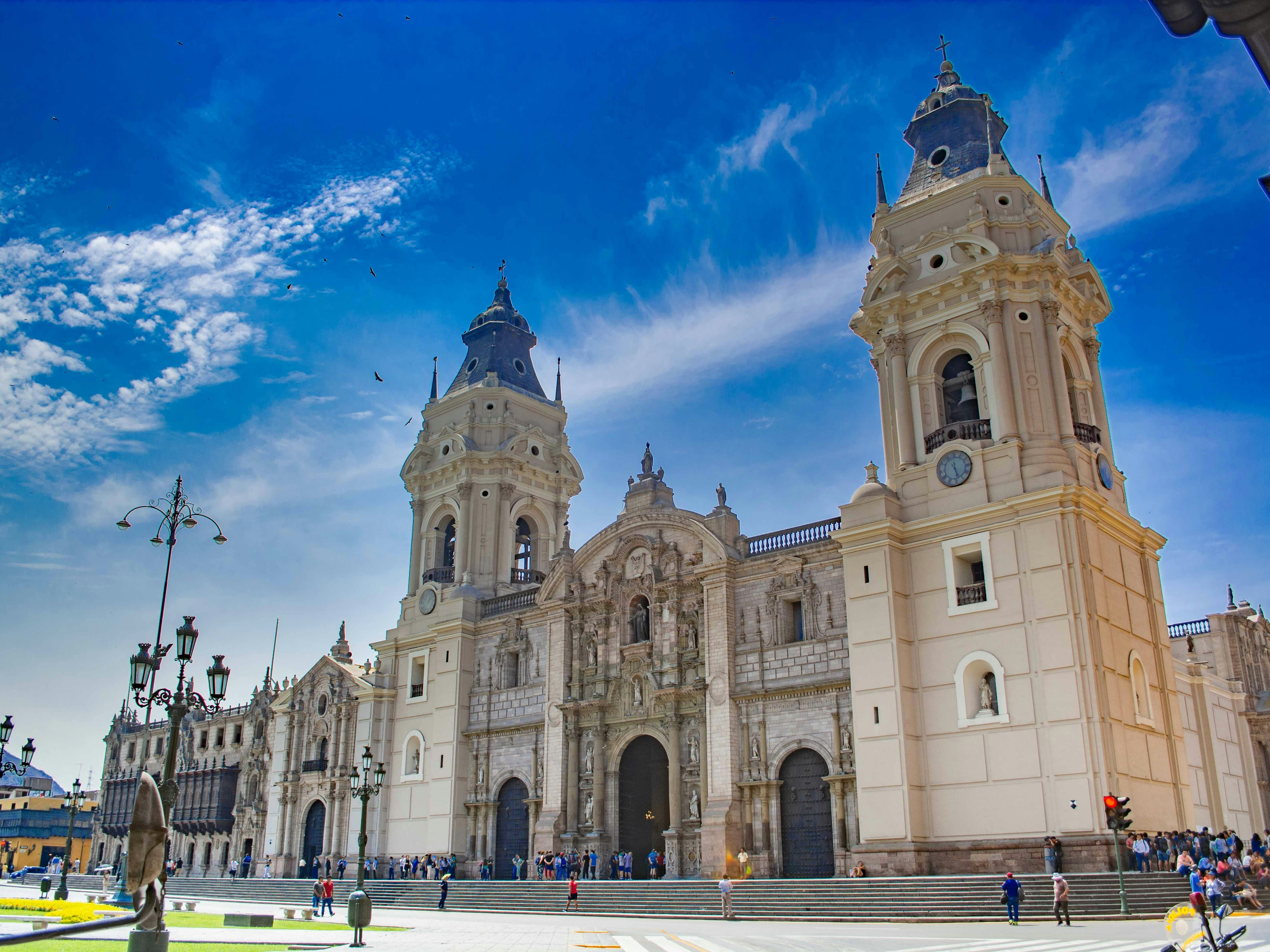 A grand cathedral with two bell towers stands under a blue sky, surrounded by street lamps and people.