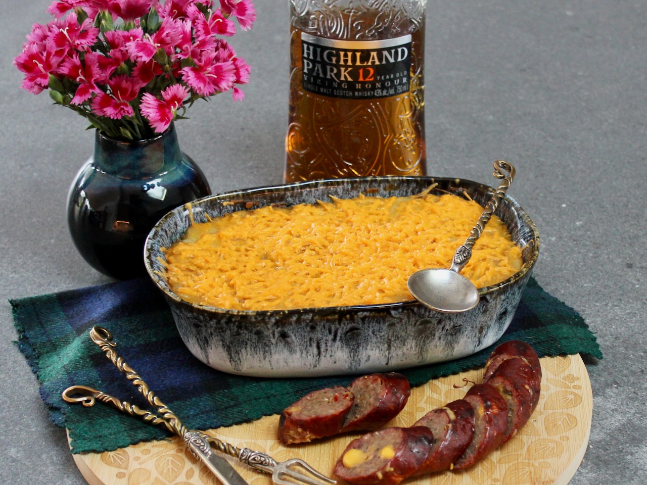 The image shows a meal setup with a casserole dish, sliced sausage on a wooden board, a bottle of Highland Park 12 whiskey, and a vase of pink flowers.