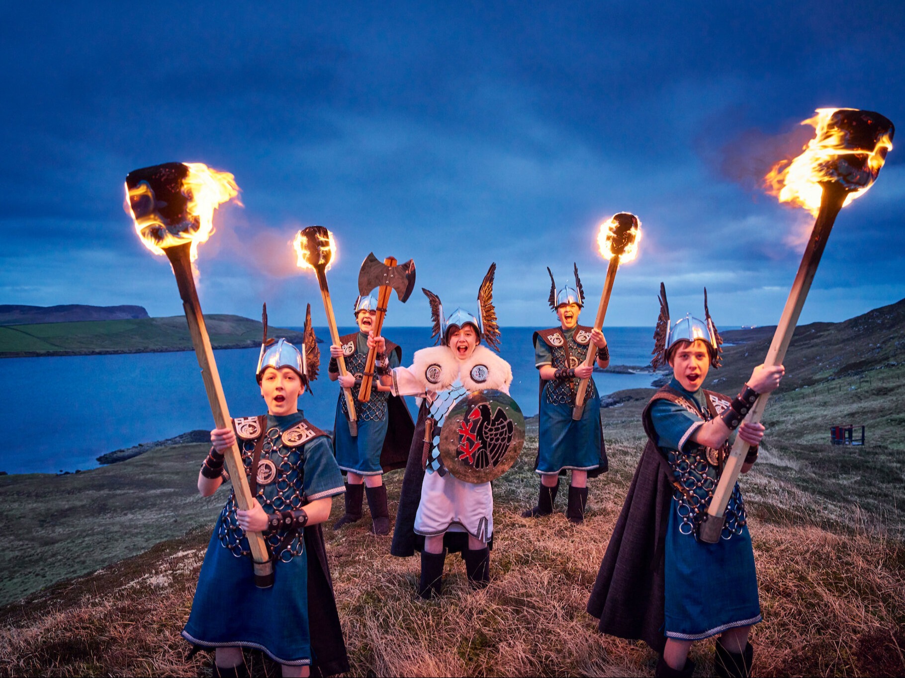 The image depicts a group of individuals dressed in historical or fantasy-inspired attire, holding flaming torches against a twilight sky with a body of water in the background.