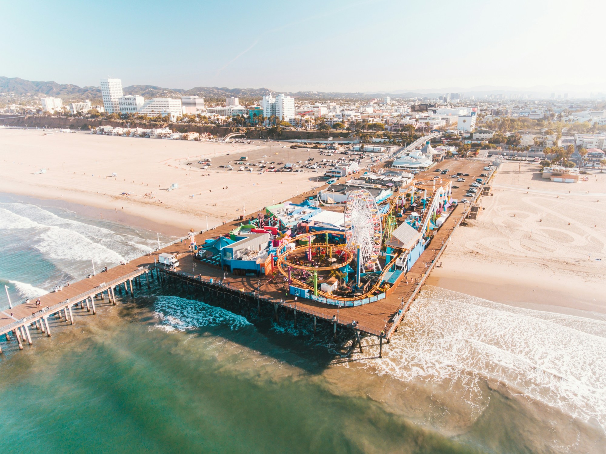 Aerial view of the beach and Santa Monica pier as the waves lap the shore on a sunny day.