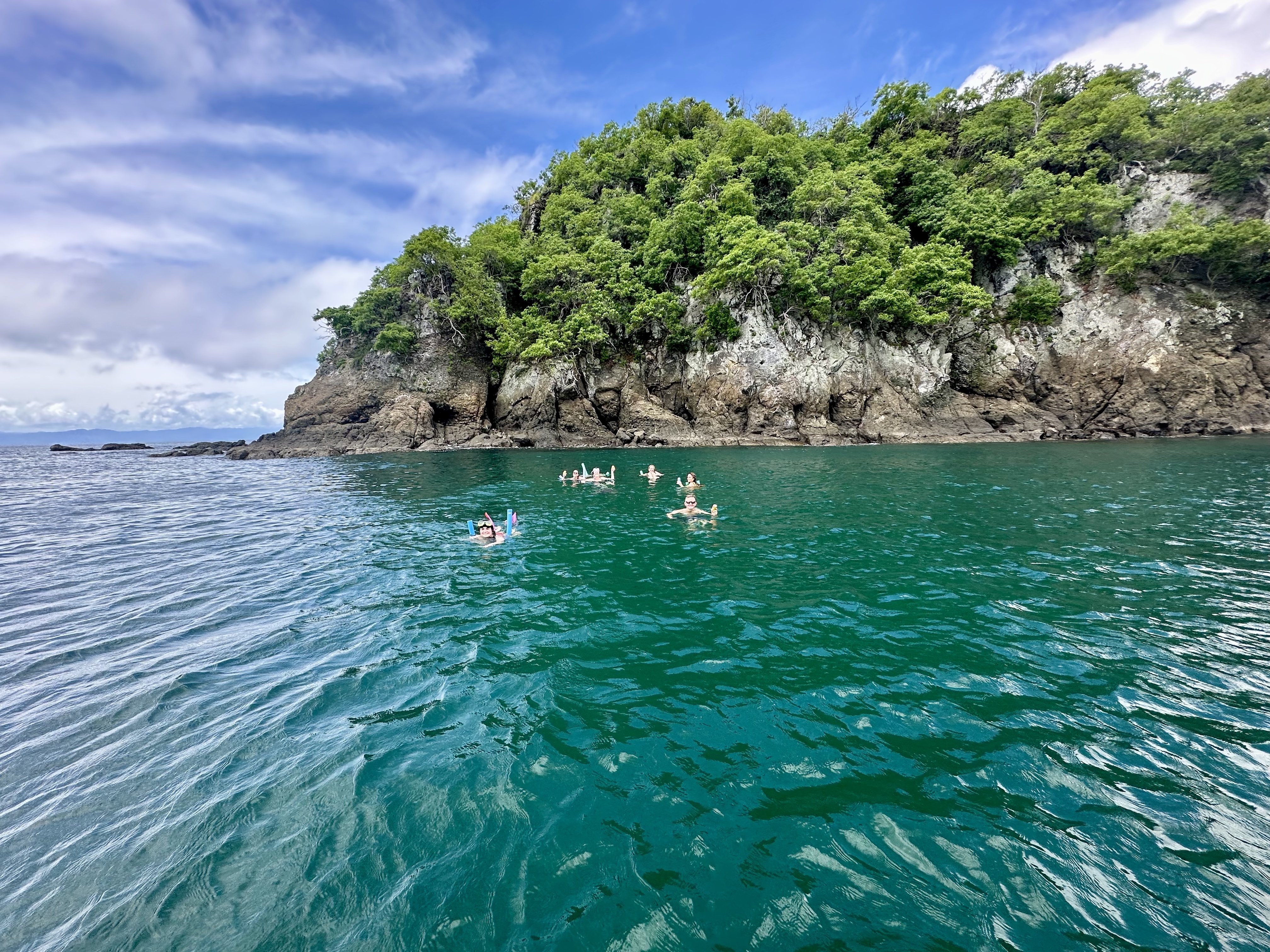 A group of individuals swimming in clear greenish-blue waters near a rocky cliff with lush green vegetation.