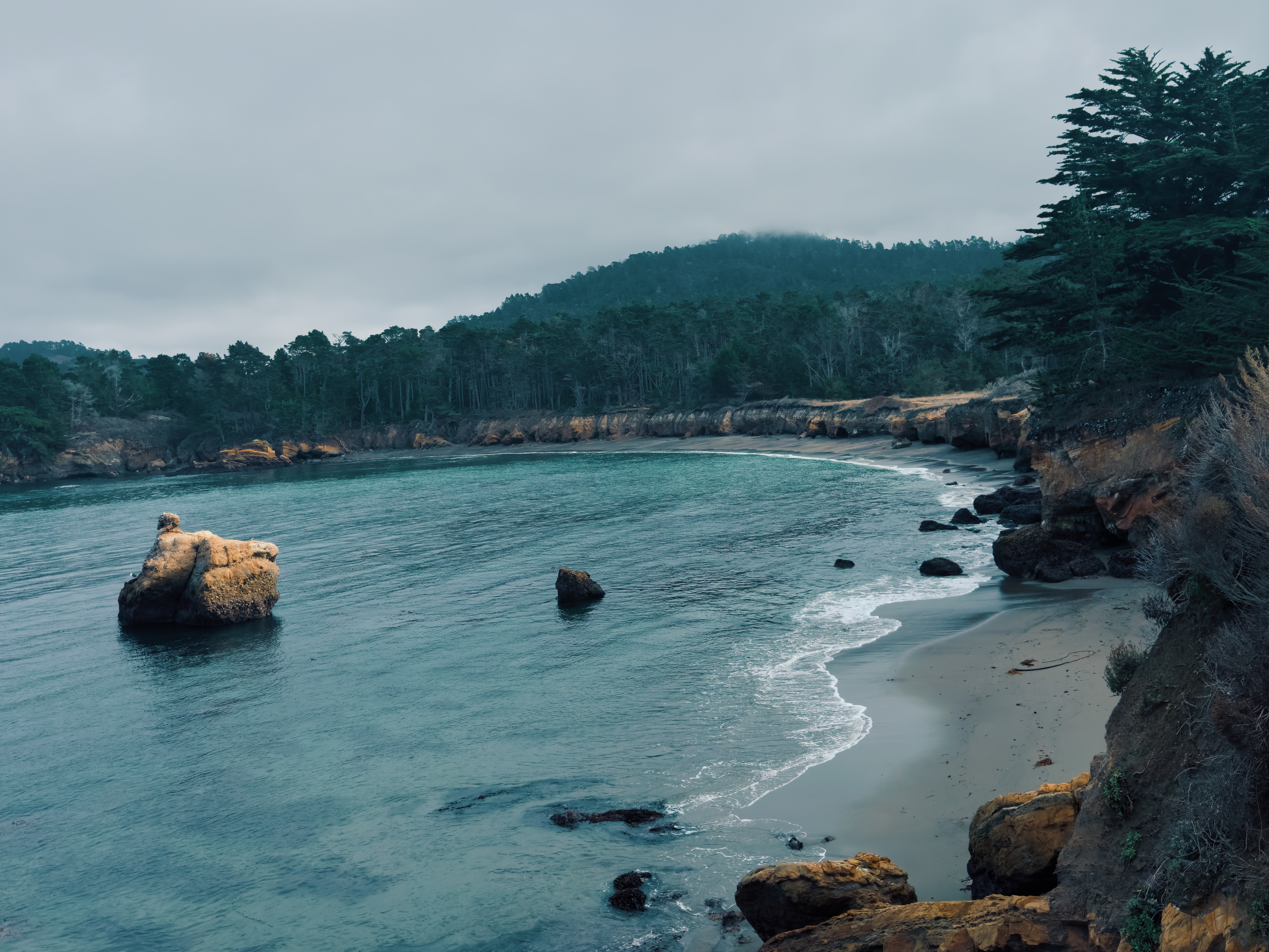 The image shows a peaceful coastal scene with a rocky shoreline, calm sea and forested hills under a cloudy sky.