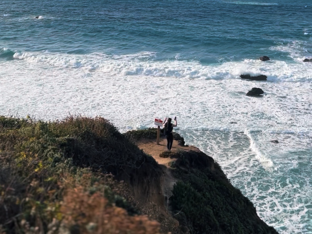 A scenic coastal landscape with waves crashing against the shore and people walking along a cliffside trail.