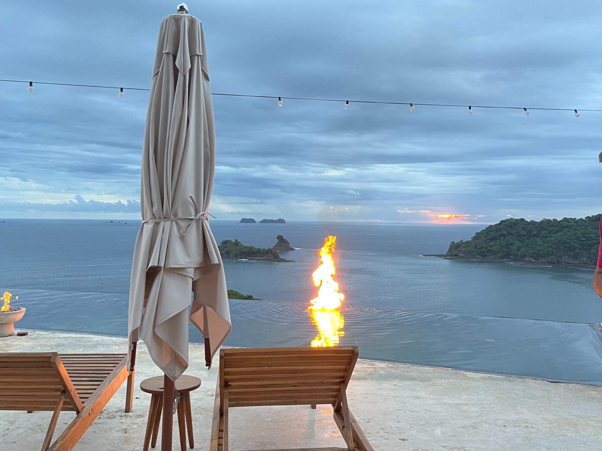 A view of the ocean from a deck with wooden chairs, an umbrella and a fire at sunset.