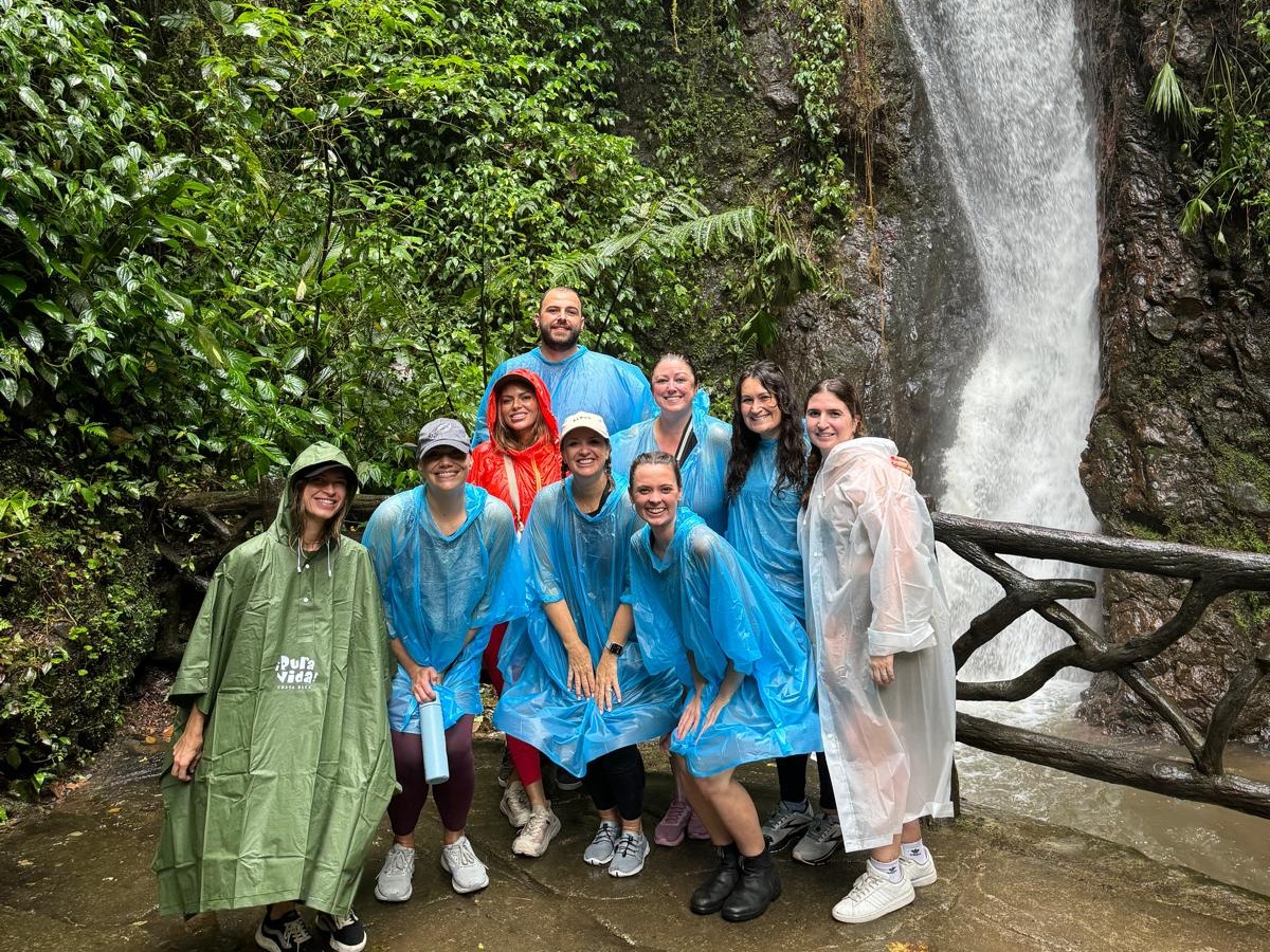 Advisor posing with a group of people wearing raincoats on a bridge in front of a waterfall and lush green forest.