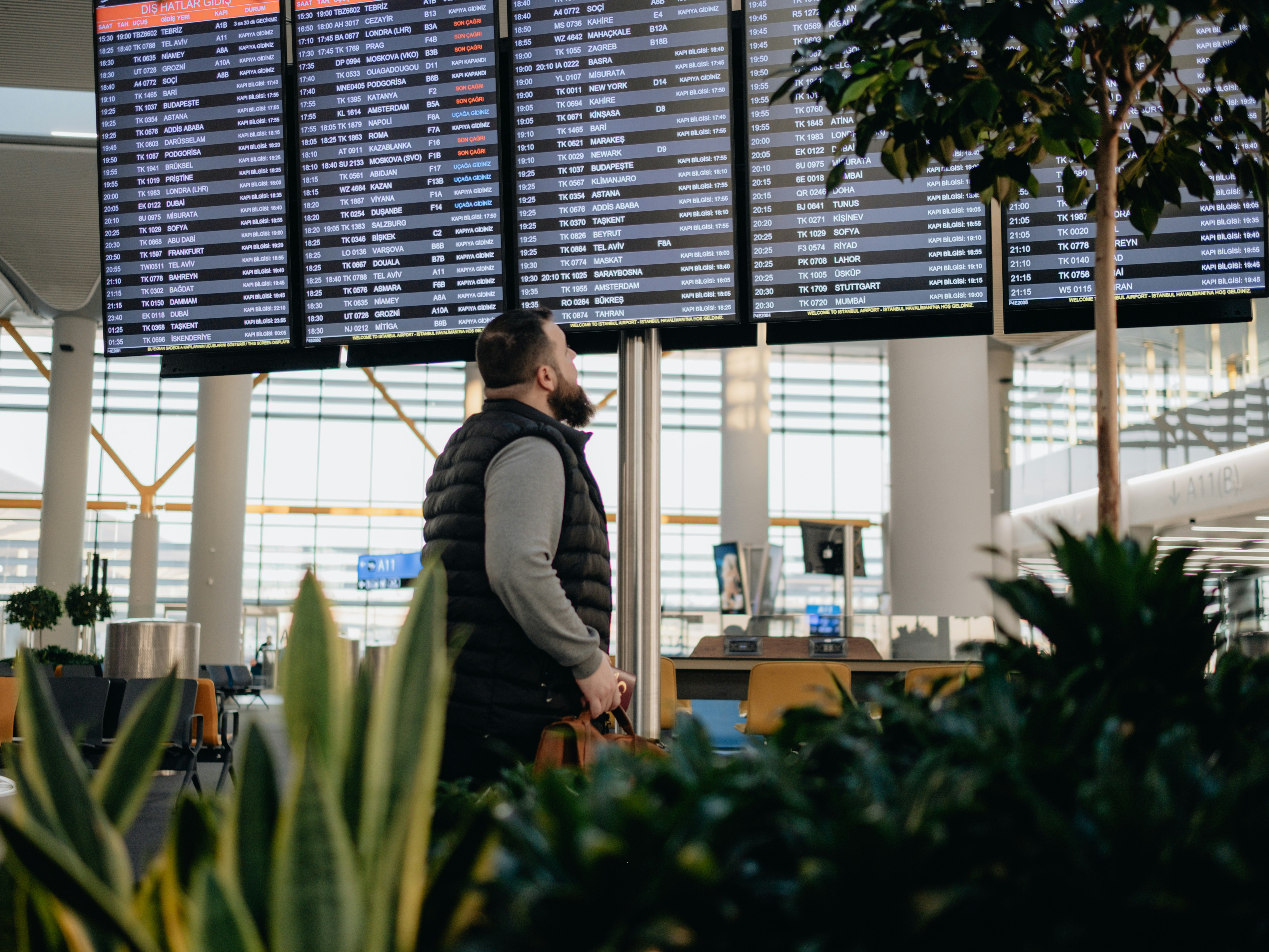 A person stands in an airport terminal, looking at a large flight information display board.