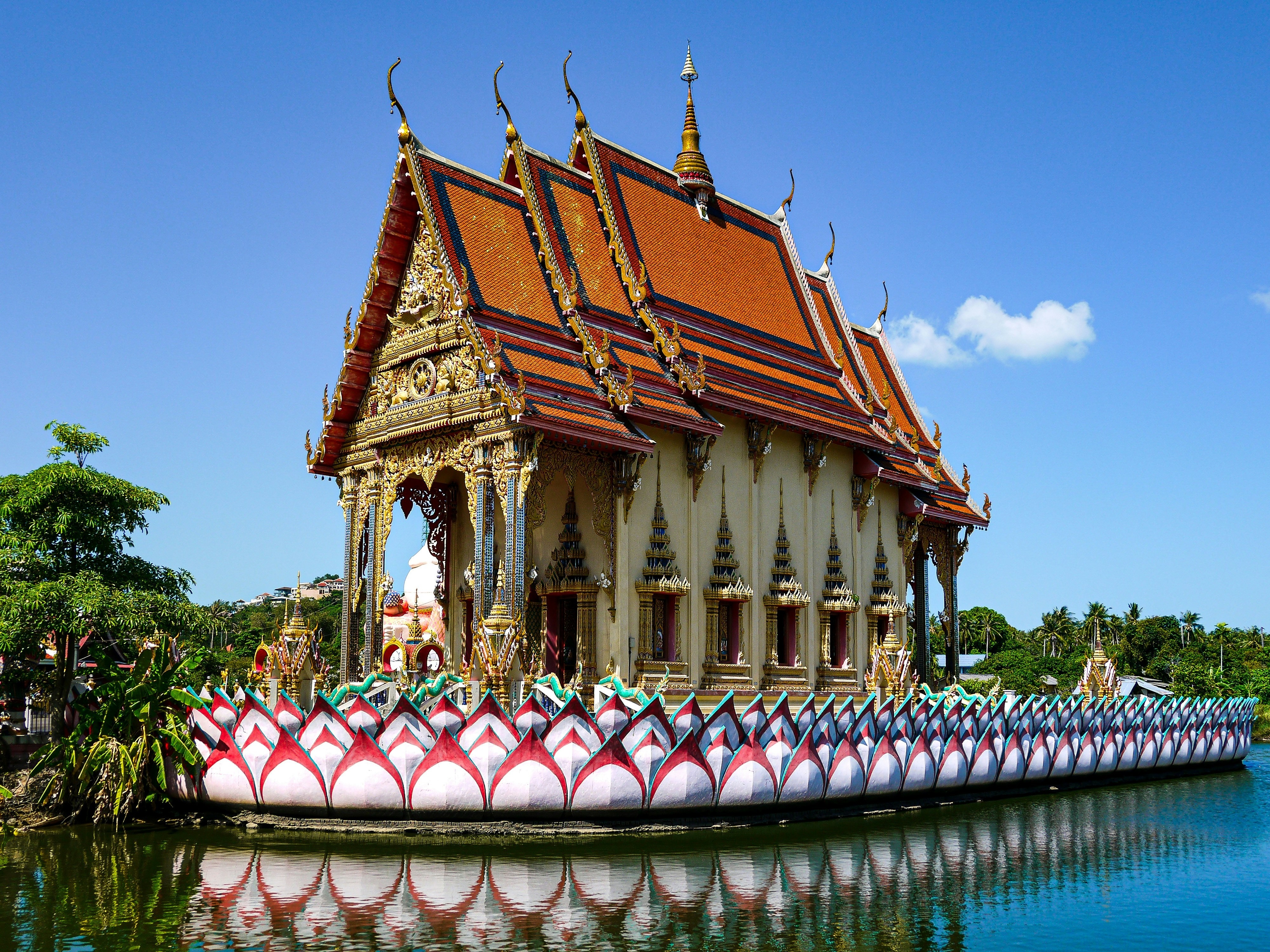 The image depicts a vibrant Thai temple with ornate roofing and decorations, surrounded by water and set against a clear blue sky.