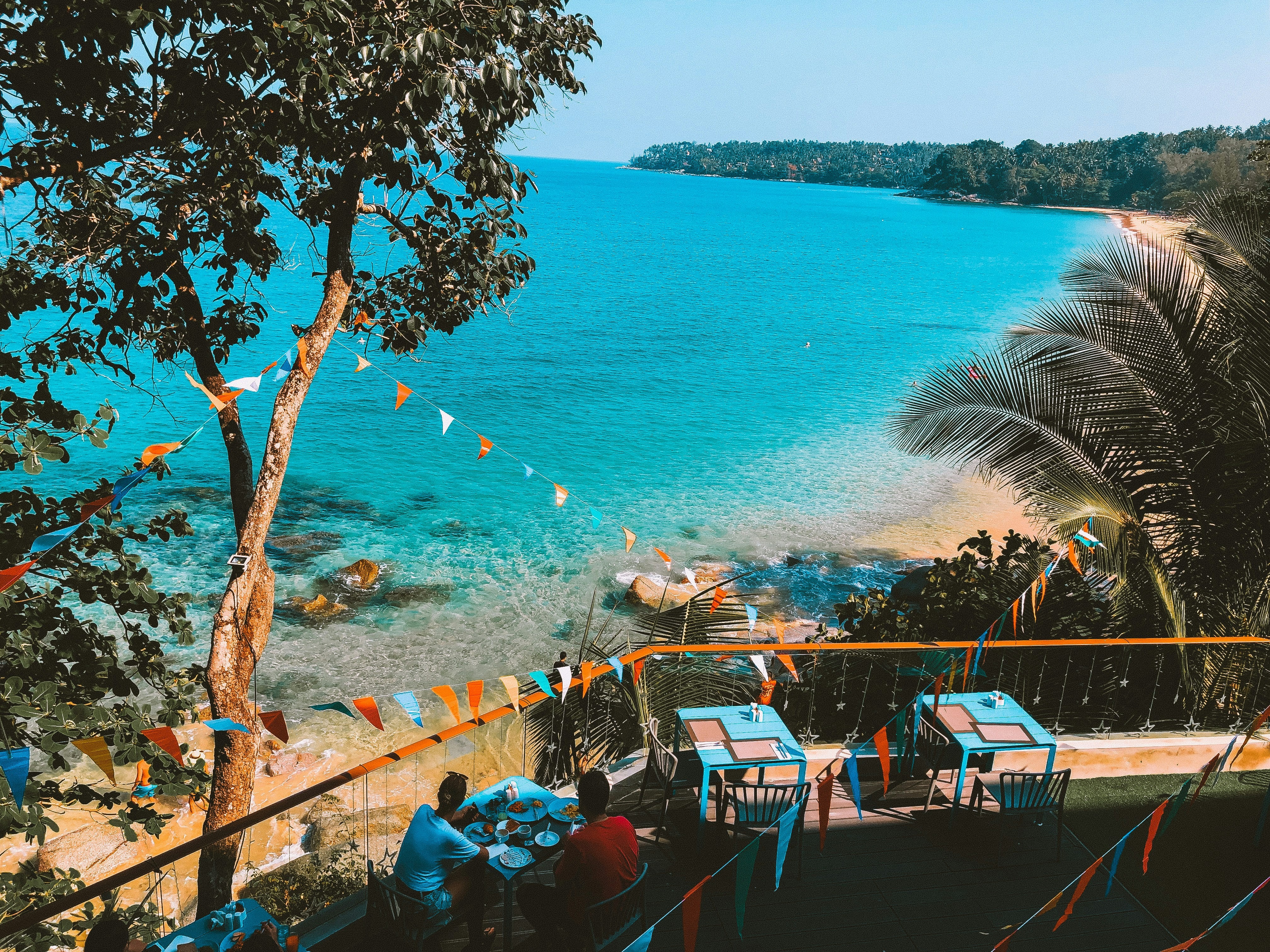 A scenic coastal view with tables set for dining near a clear blue sea, framed by lush greenery and decorative flags.