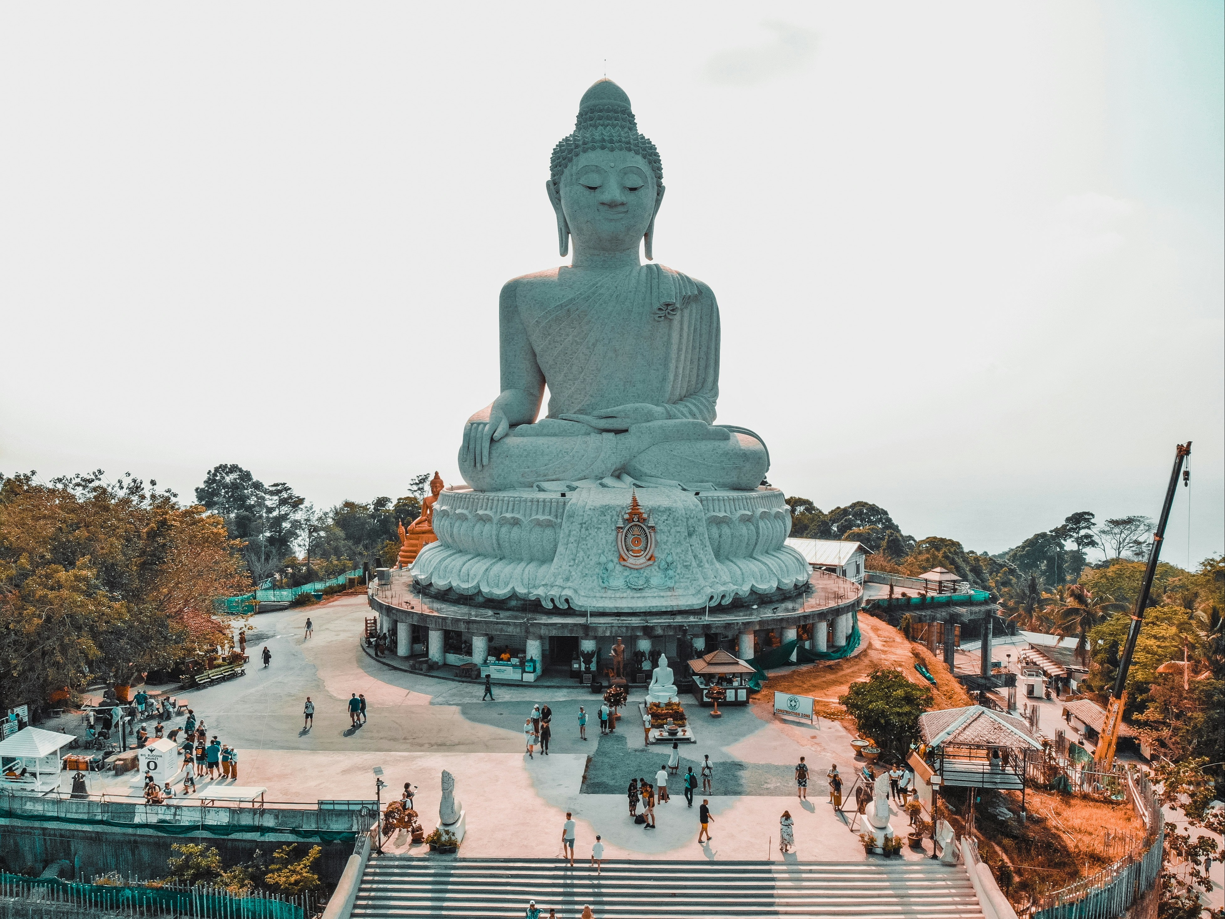 A large Buddha statue overlooks a gathering of people in a serene environment with trees and clear skies.