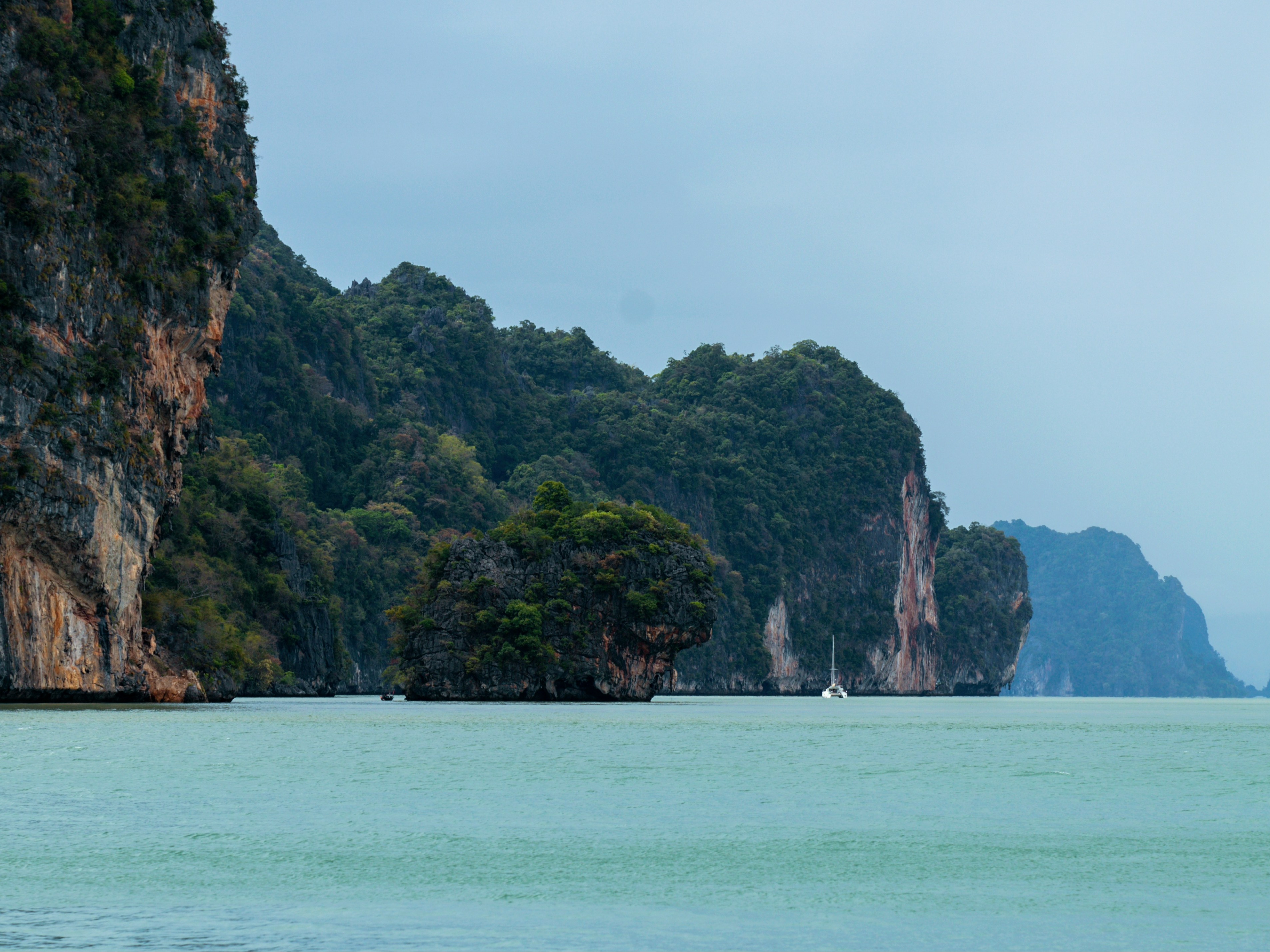 A serene seascape with towering limestone cliffs and lush greenery under a calm, overcast sky.