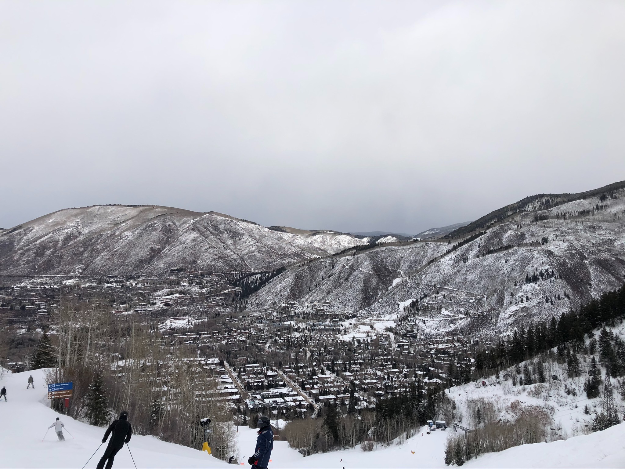 A snowy mountain landscape with skiers in the foreground and a town nestled in the valley below on a cloudy day.