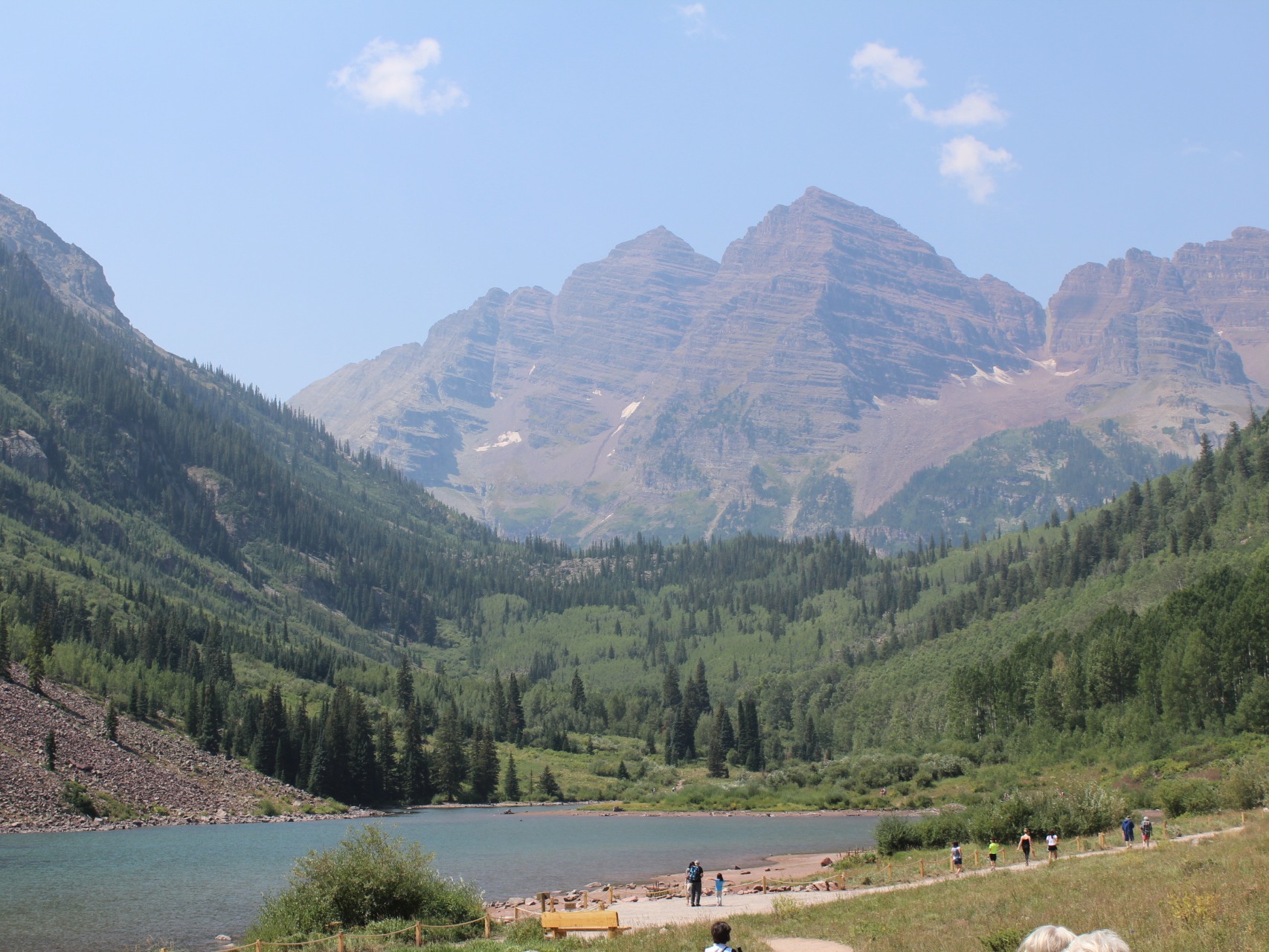 The image shows a scenic view of a mountain range with a lake in the foreground and people on a trail to the right.
