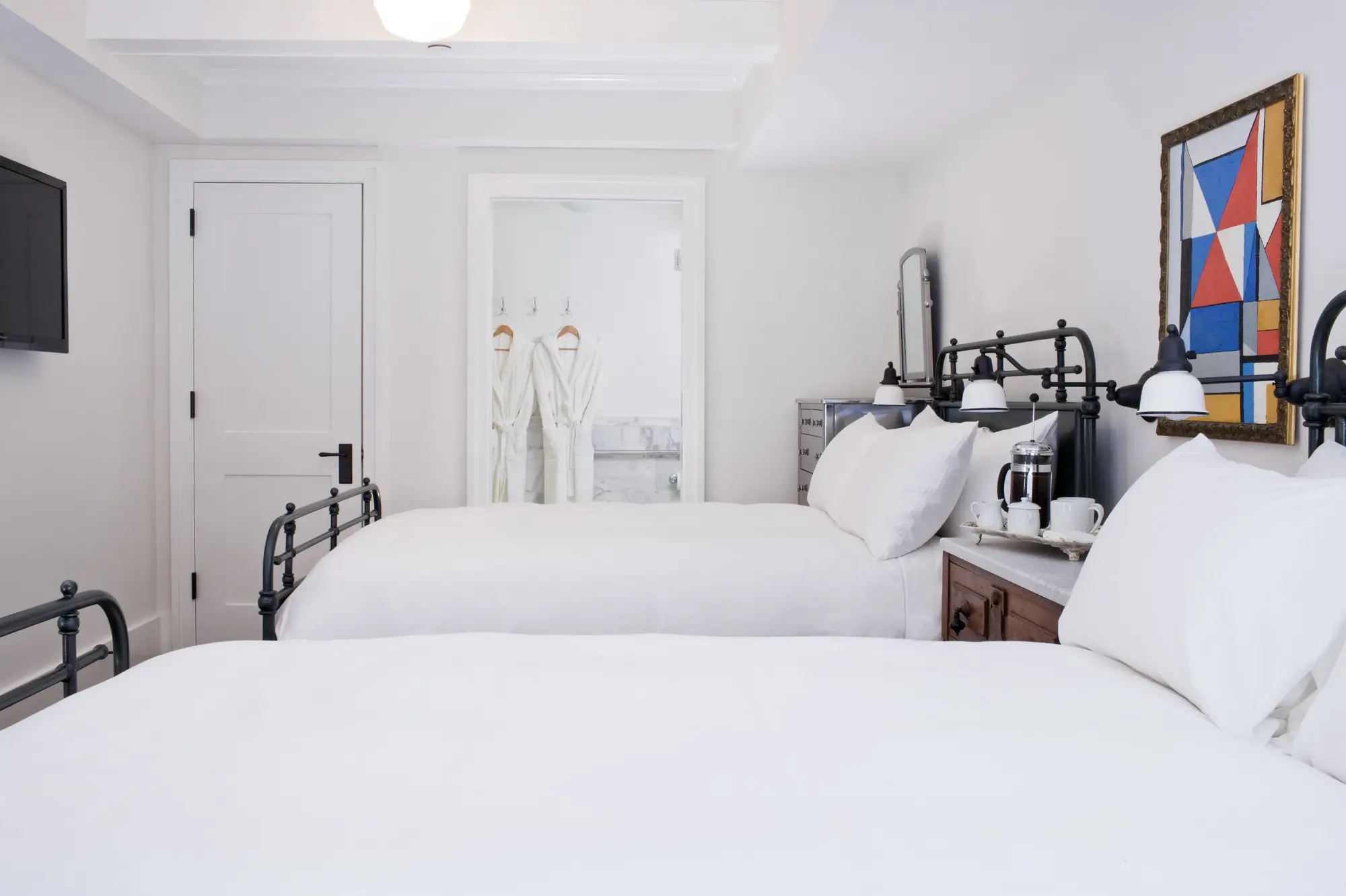 Twin beds fill a cozy, whitewashed room. Abstract wall art and contemporary decor, plus a French press and coffee cups are present, too