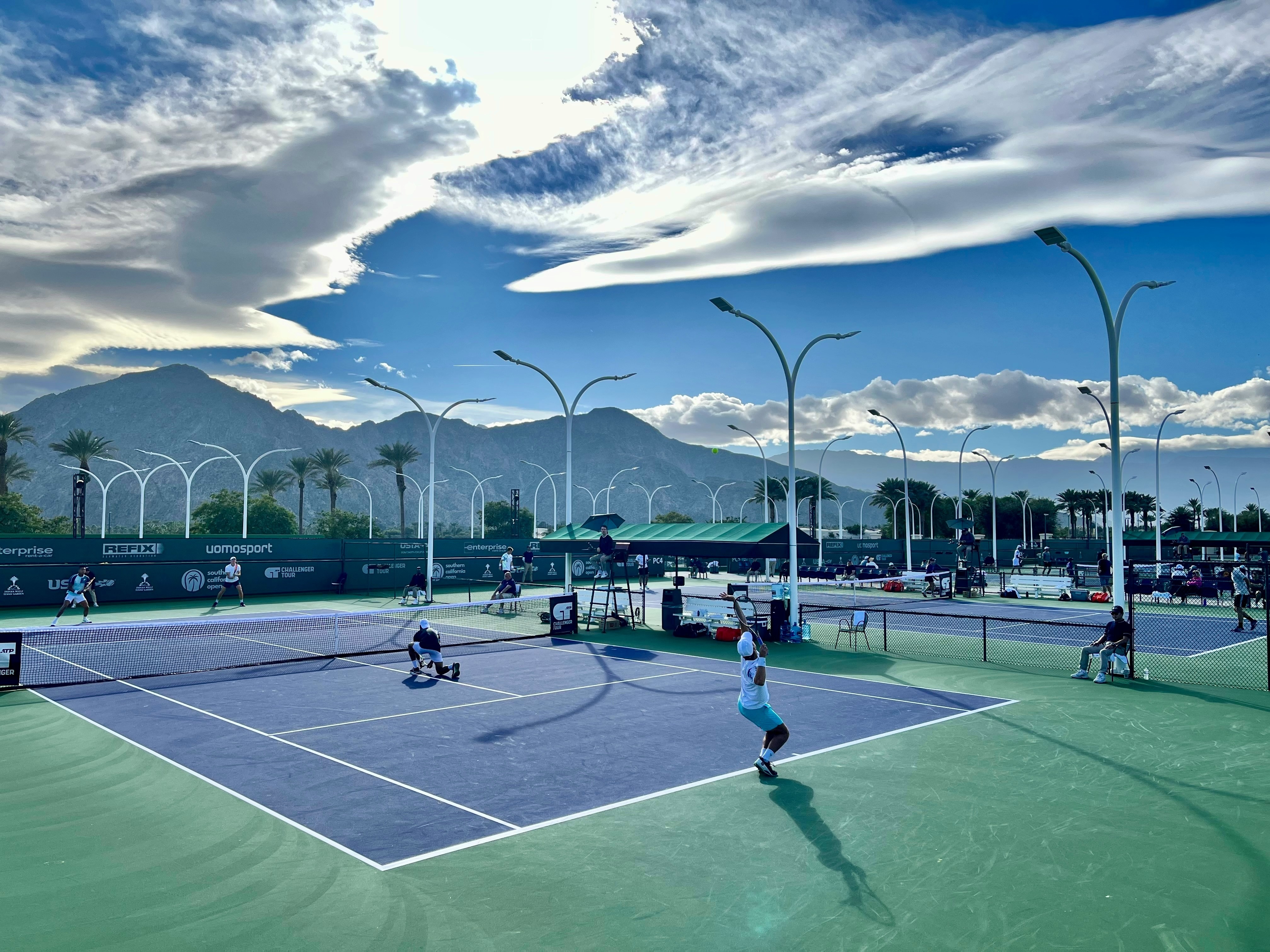 A tennis court with players in action, set against a backdrop of mountains and a dramatic sky.