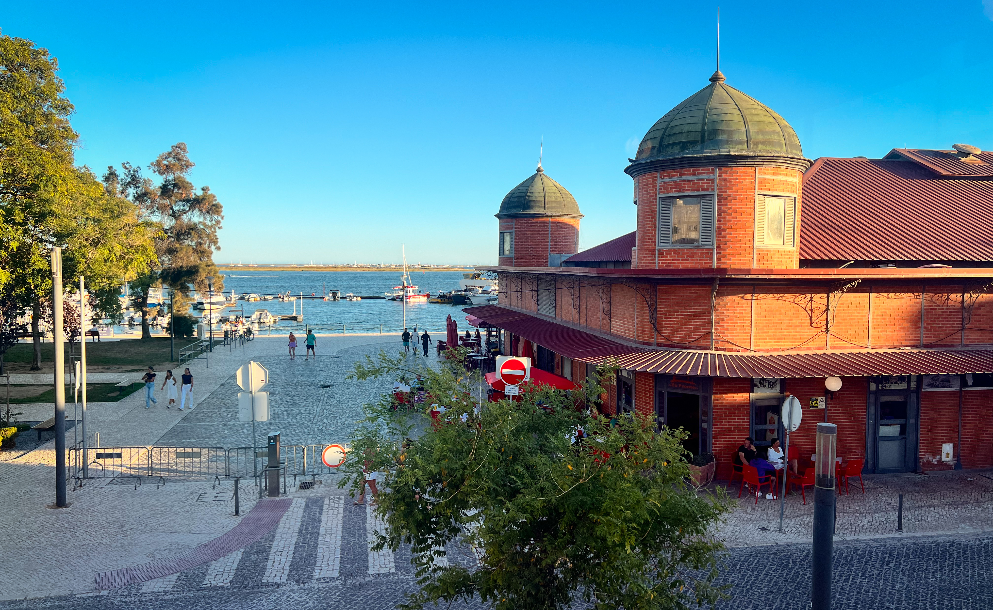 A bustling street scene at dusk with people dining at outdoor restaurants and walking along the pedestrian road flanked by traditional white buildings with terra cotta roofs.
