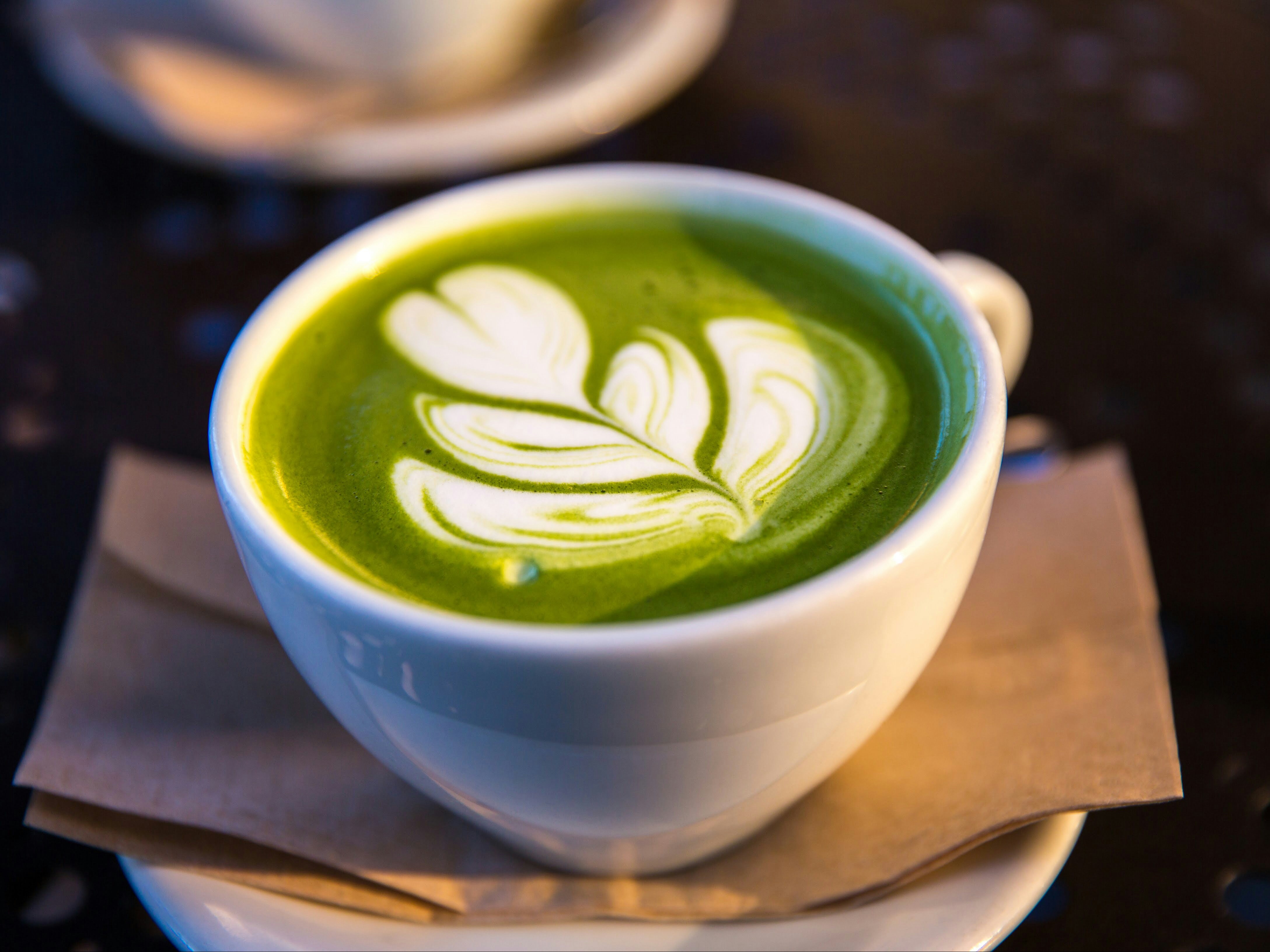 A close-up of a cup of green matcha latte with a detailed white leaf-like art design on its surface, placed on a saucer over a napkin.
