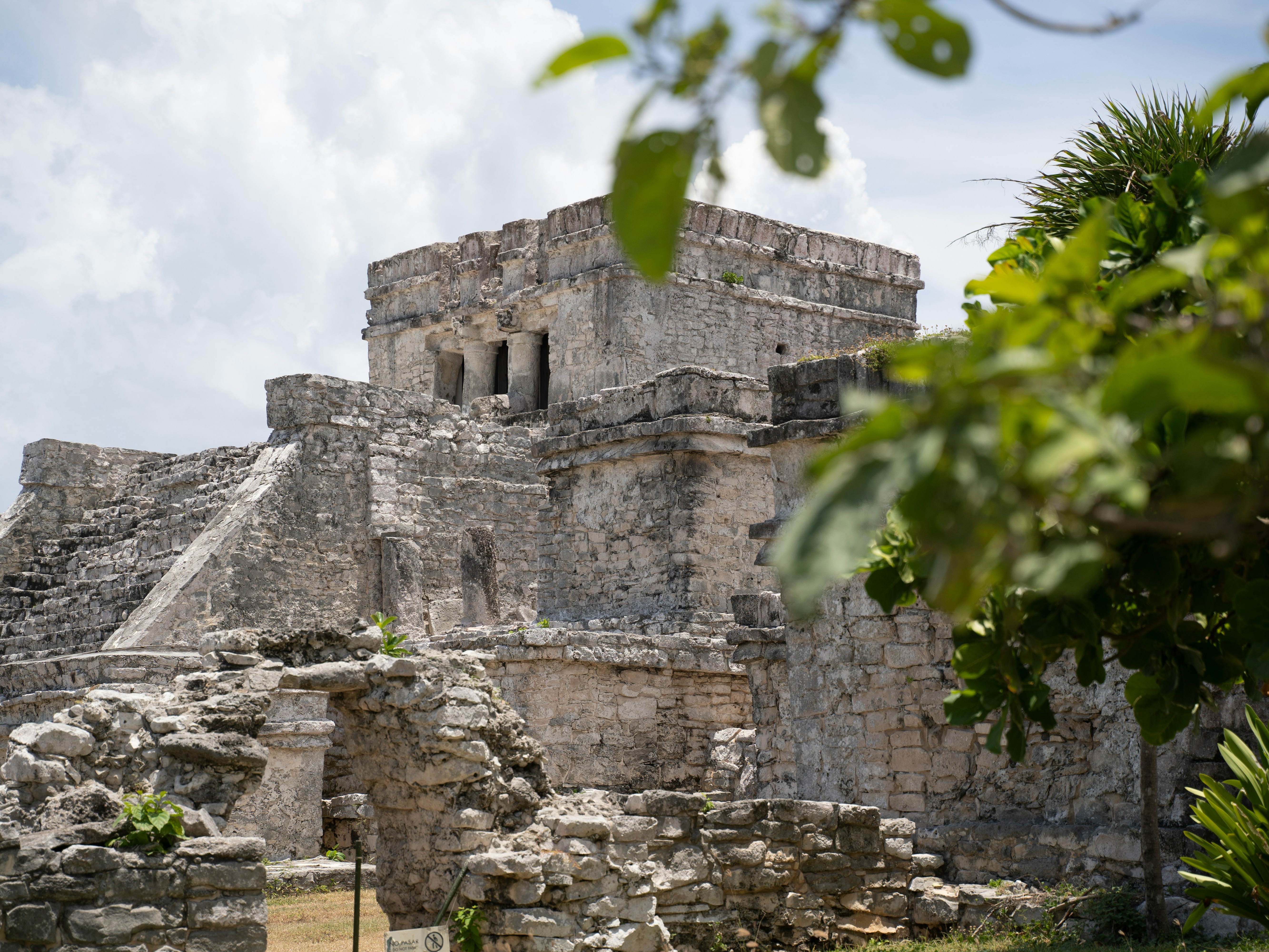 An ancient stone structure, possibly Mayan ruins, with a clear sky and green foliage in the background.