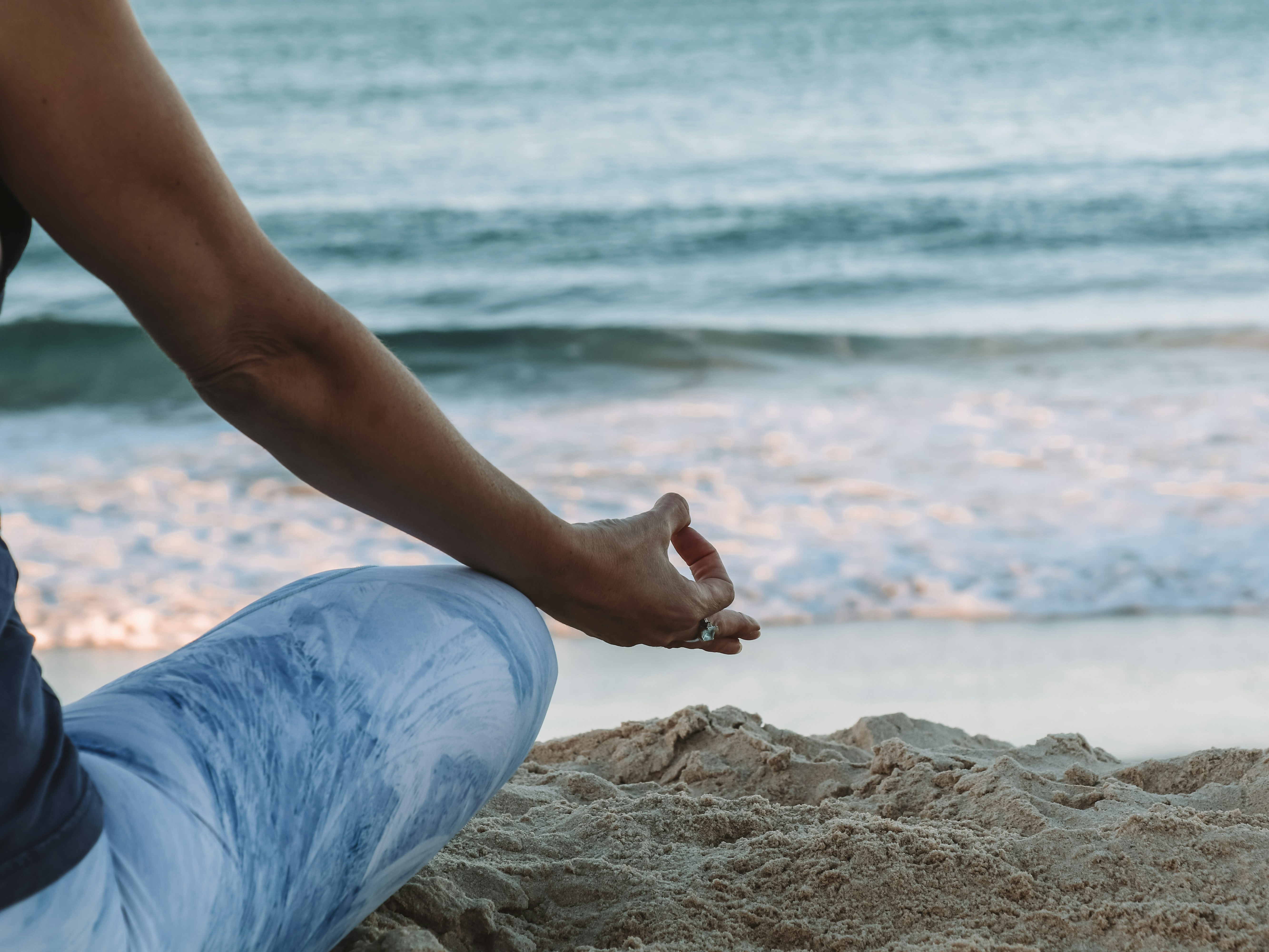 A close-up of a person’s side, seated on the beach with their hand resting on the sand and the ocean in the background.