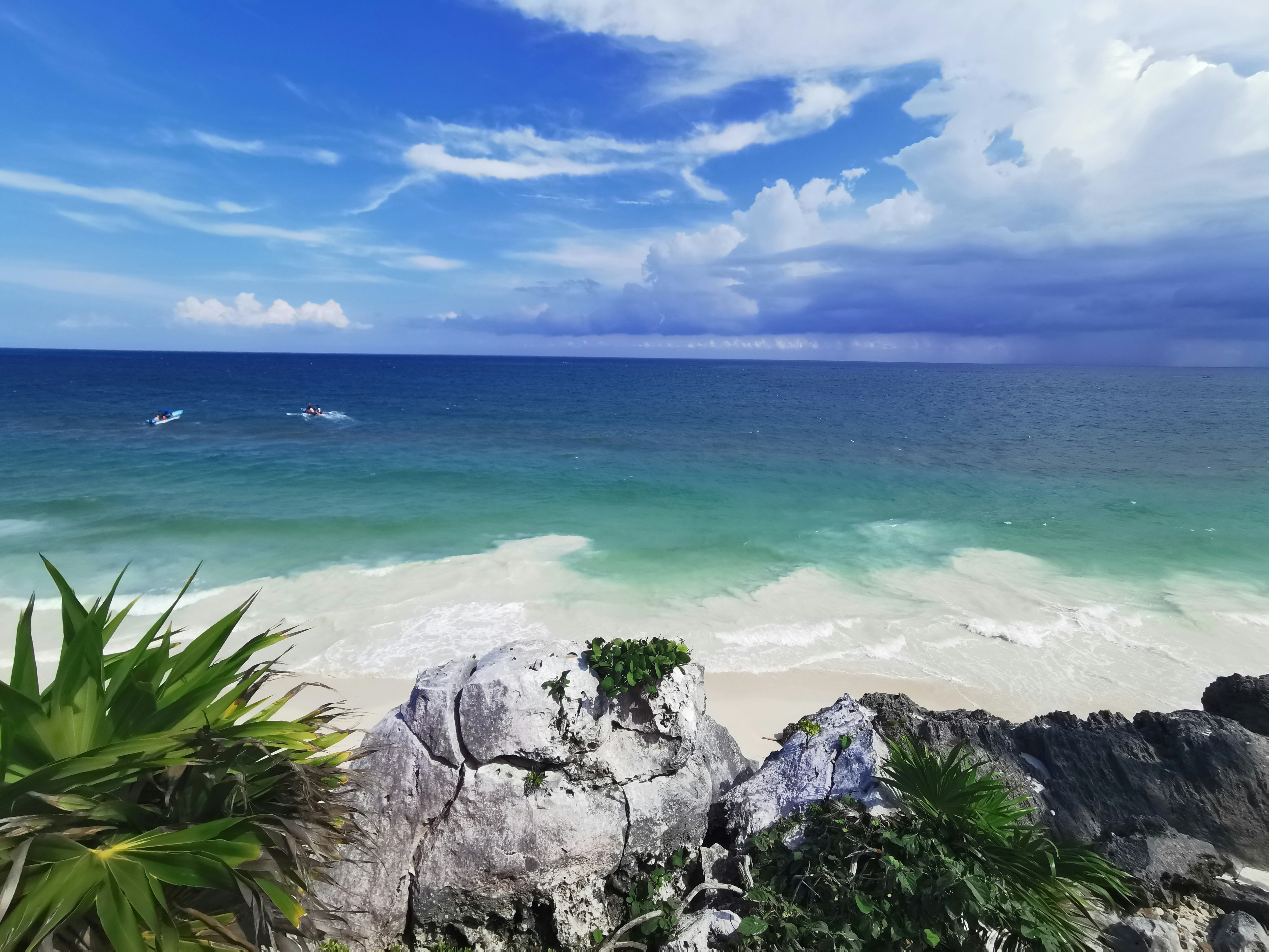 A scenic beach view with clear blue skies, turquoise sea, white sand and a rocky outcrop with green foliage.