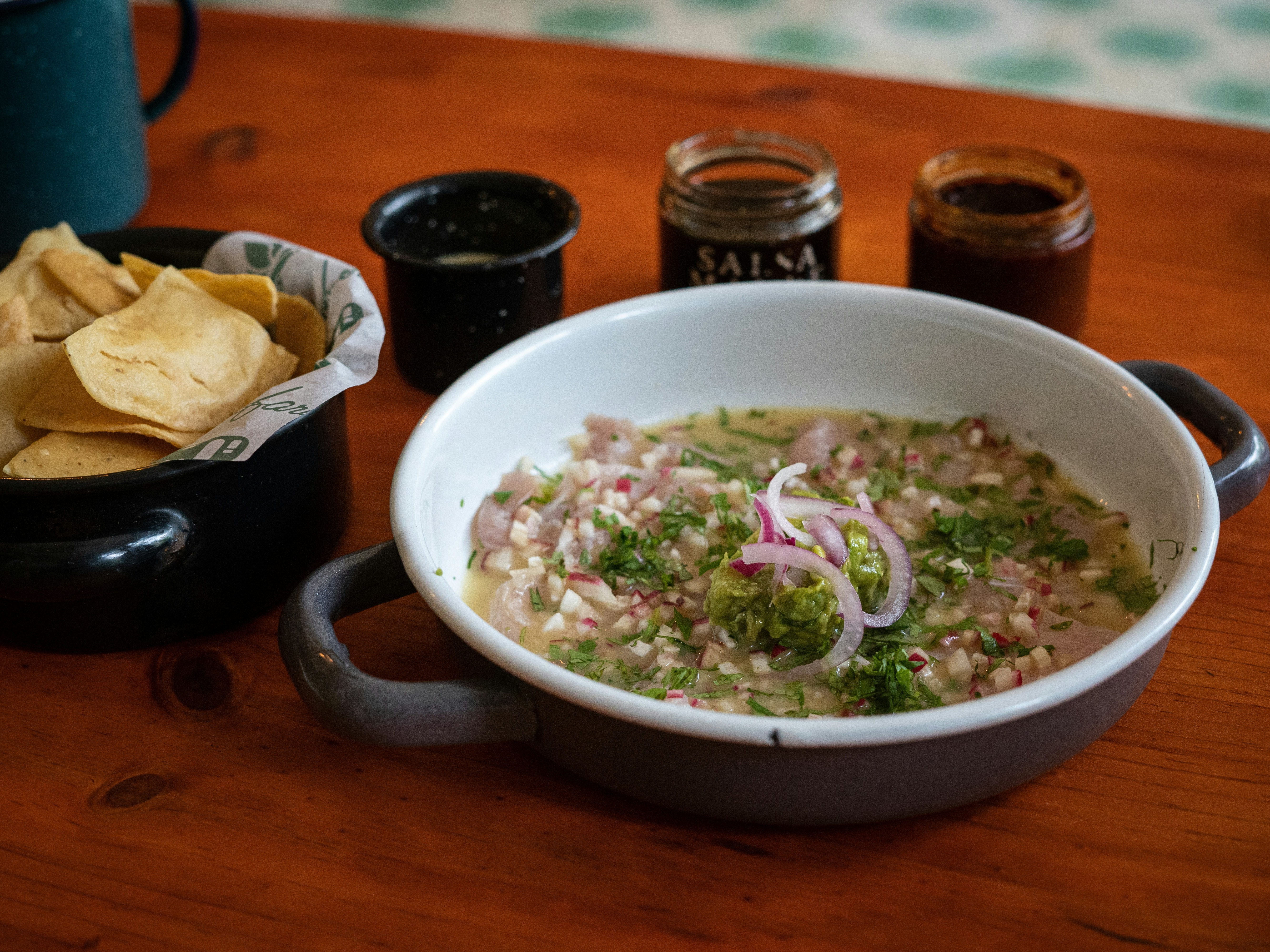 A bowl of soup garnished with herbs and onions, accompanied by tortilla chips and condiments on a wooden table.