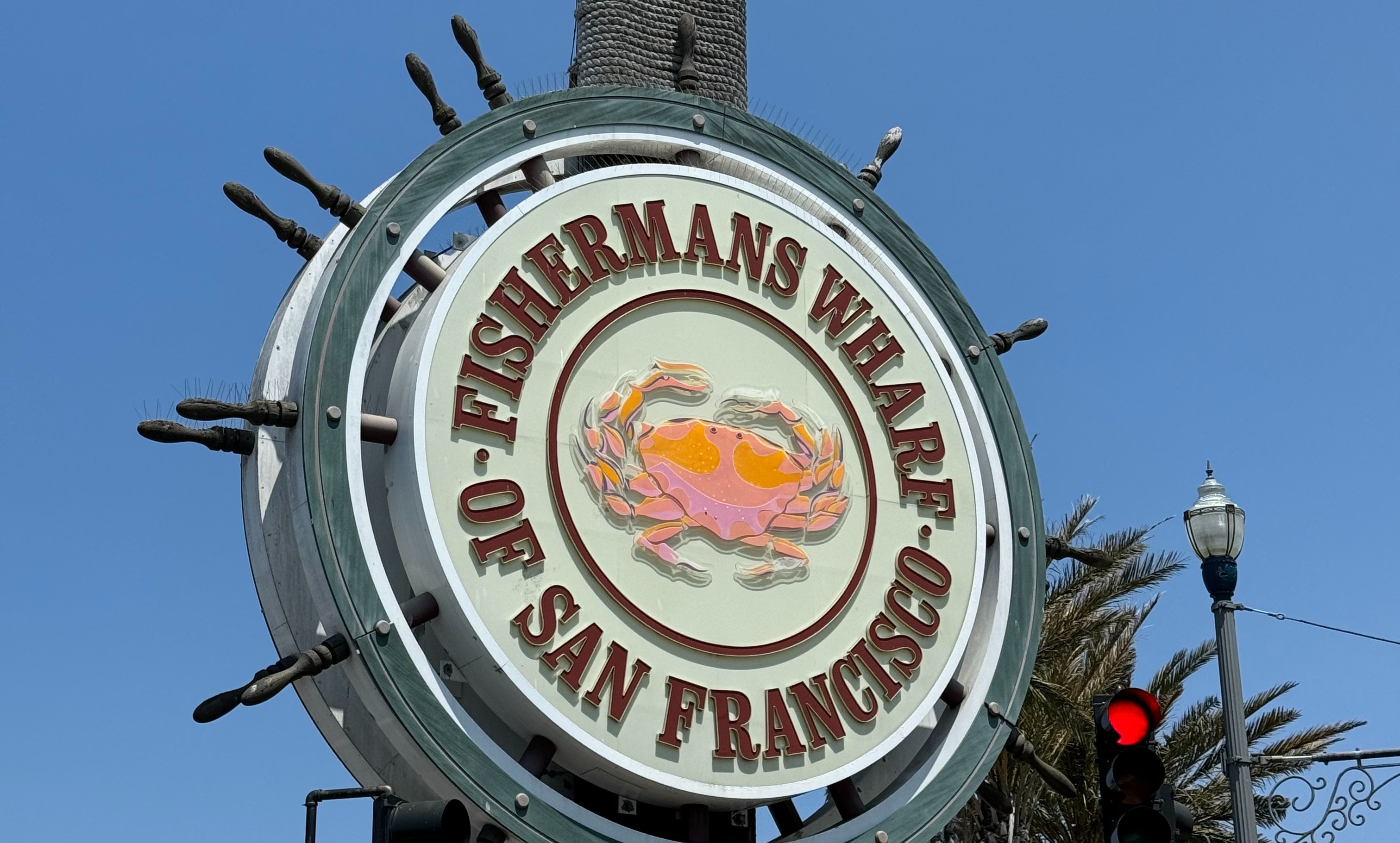 A large circular sign reads “Fisherman’s Wharf San Francisco” with a crab illustration in the center, set against a clear blue sky.