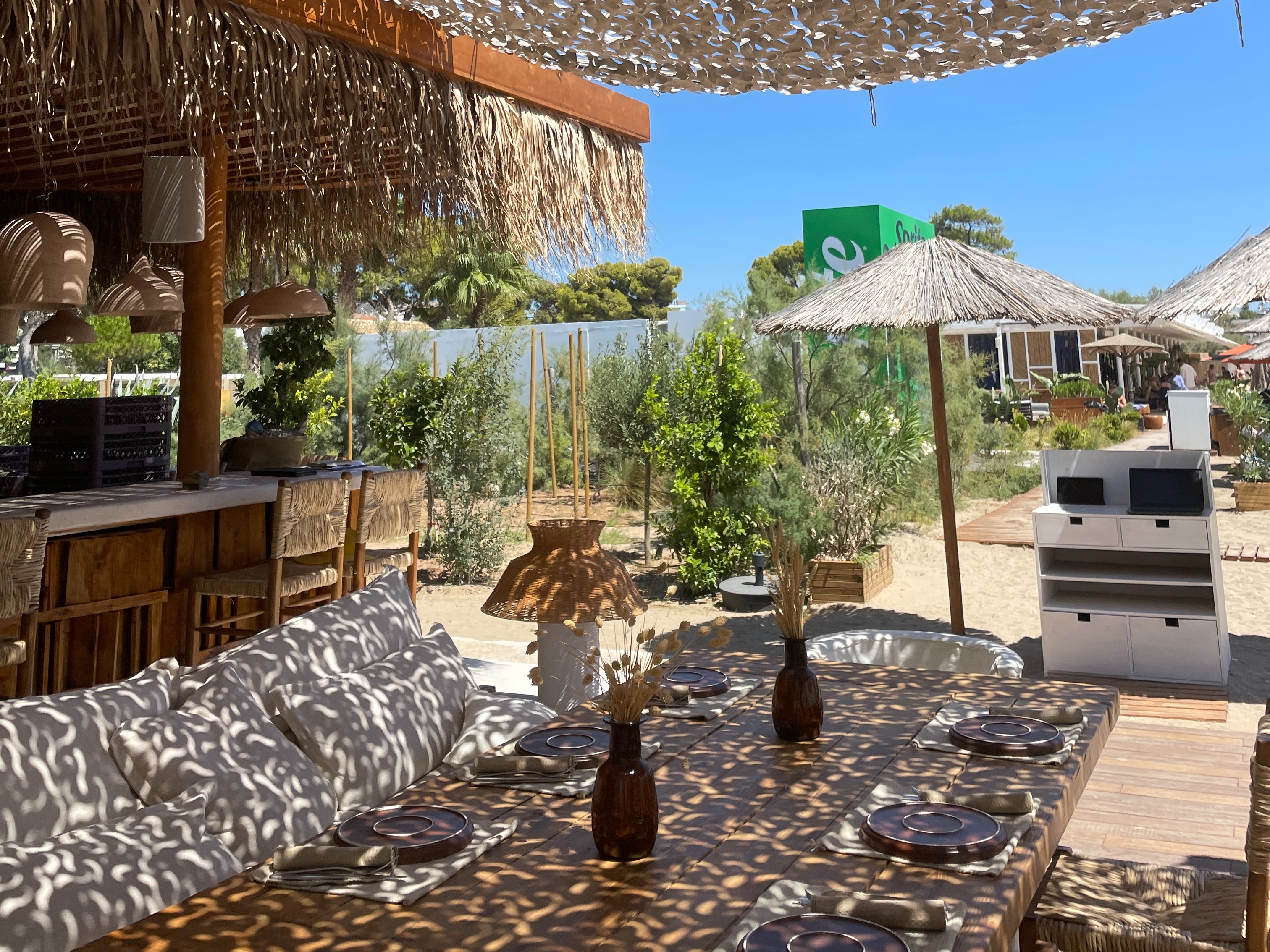 An outdoor seating area with tables and chairs, covered by a unique canopy of numerous small, reflective elements, under a clear blue sky.